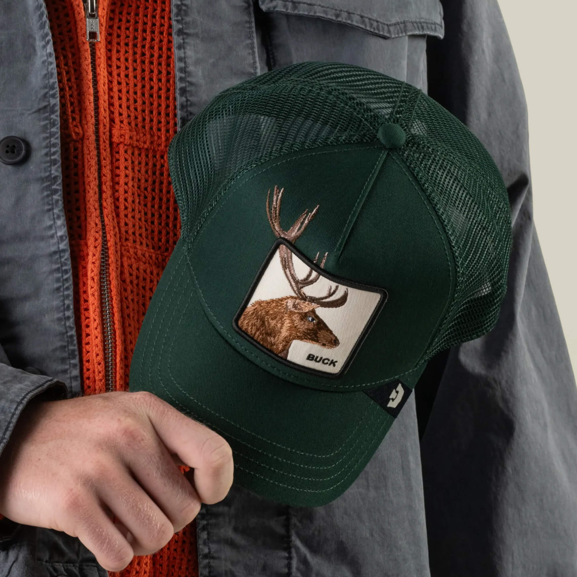 A person holds the Goorin Bros. Over the Line Buck dark green trucker hat, featuring an embroidered patch with a buck and antlers and breathable mesh panels, while wearing an orange sweater and a gray jacket.