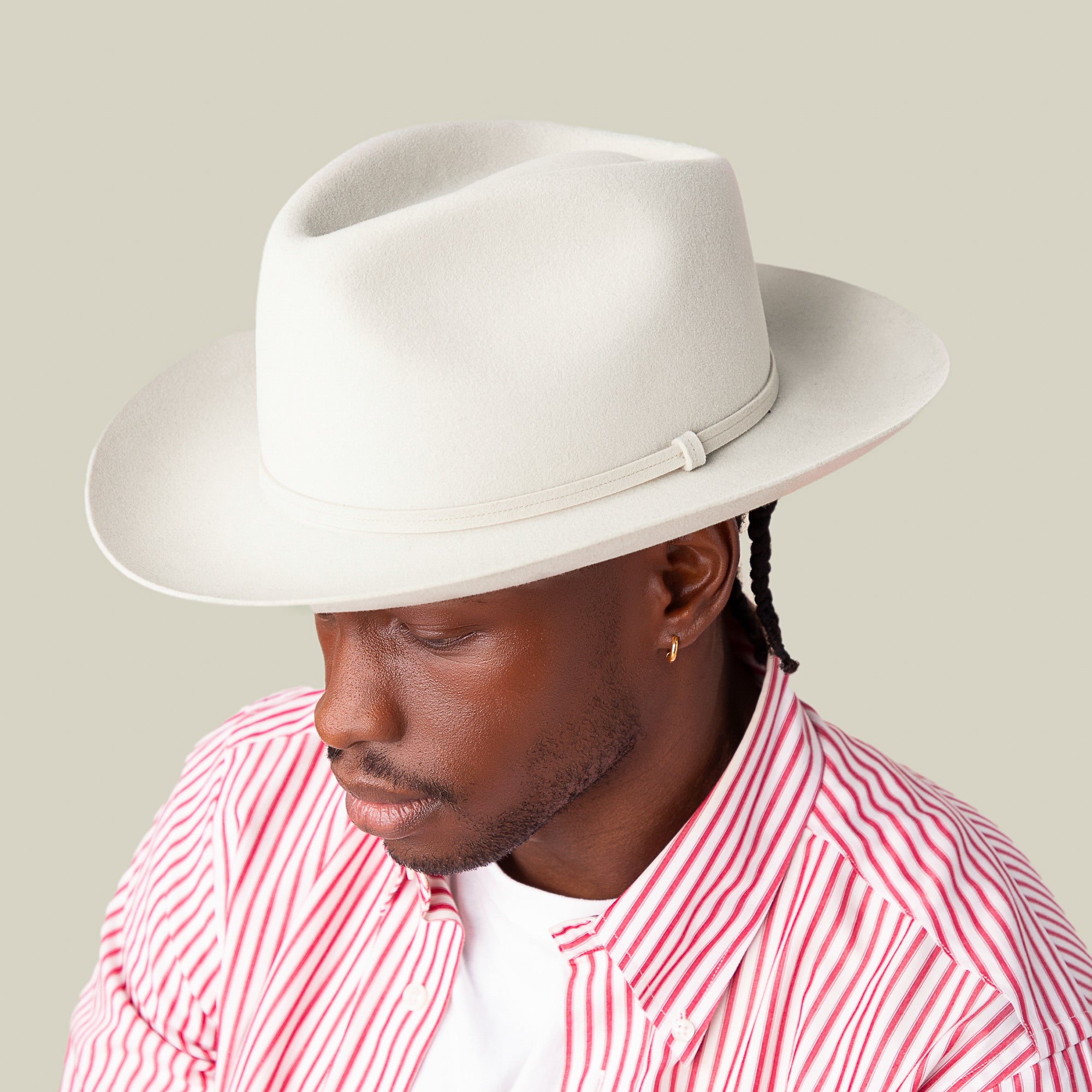 A person in a red-and-white striped shirt wears the Goorin Bros. Night Hawk, a light-colored teardrop crown fedora with a wide snap brim, while looking downward against a plain beige background.