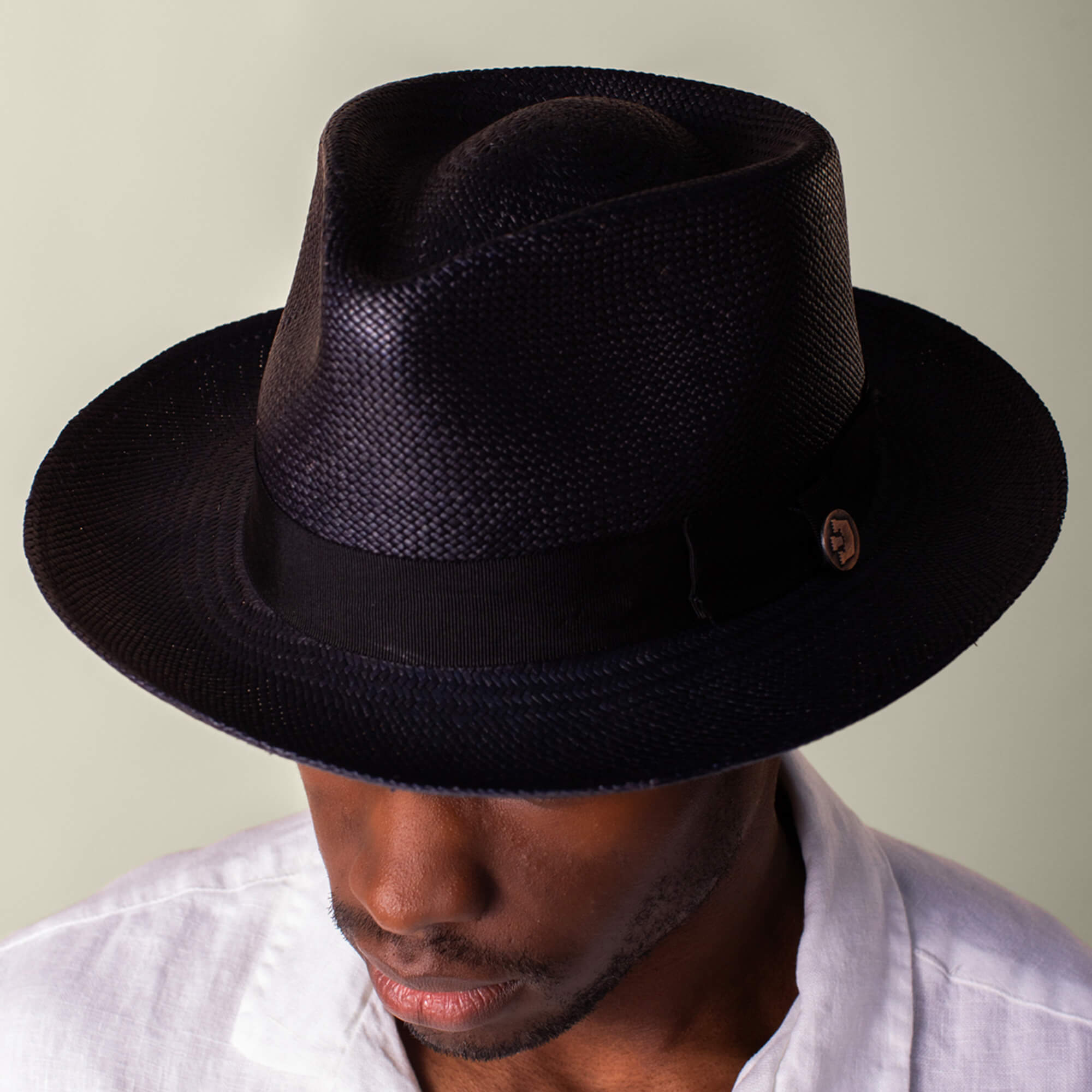 A man wears the Goorin Bros. John Sr., a black toquilla fedora hat with 50 UPF sun protection, and a white shirt, looking downward against a neutral background.