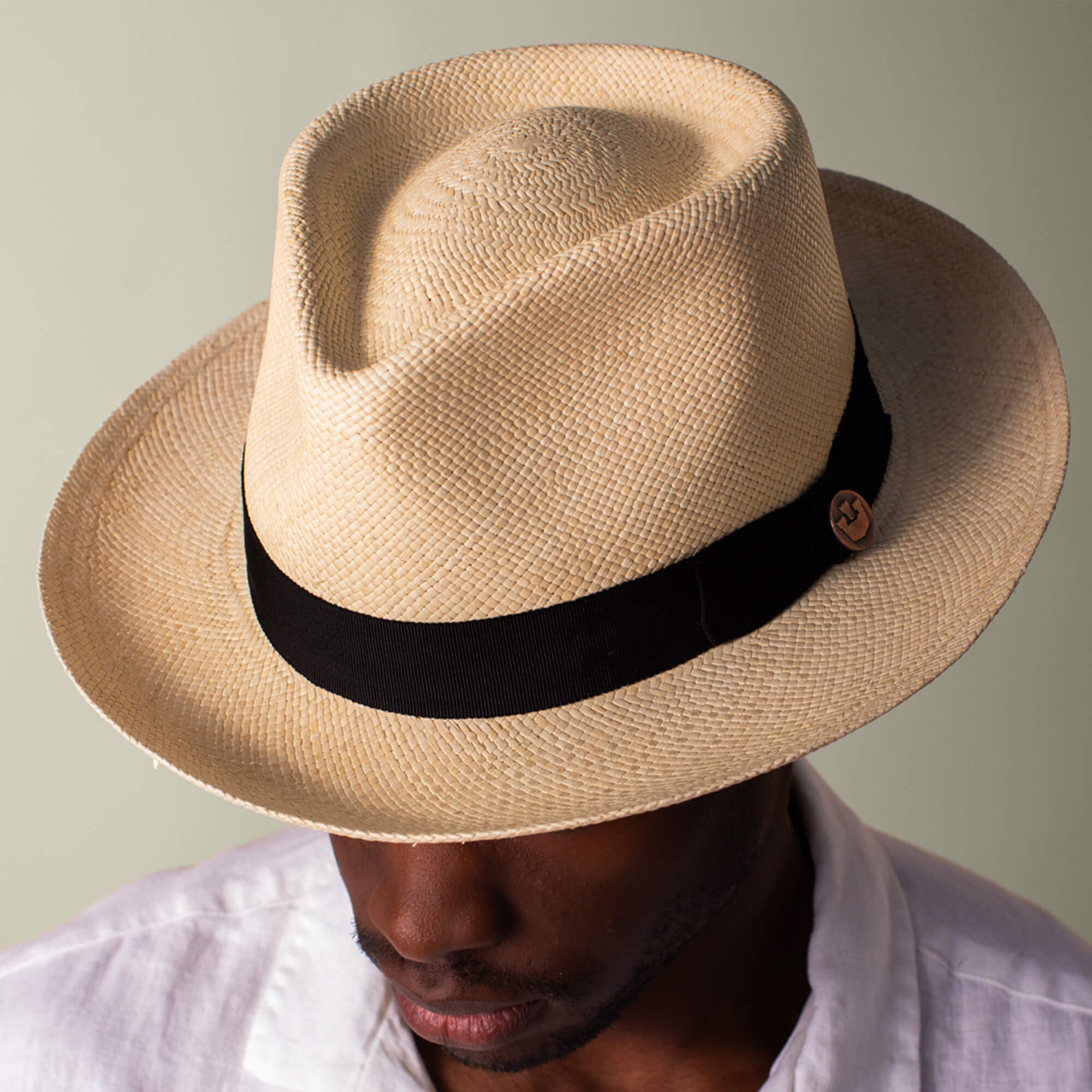 A person in a Goorin Bros. John Sr. light-colored toquilla teardrop fedora with black band (50 UPF), paired with a white shirt and looking slightly downward.