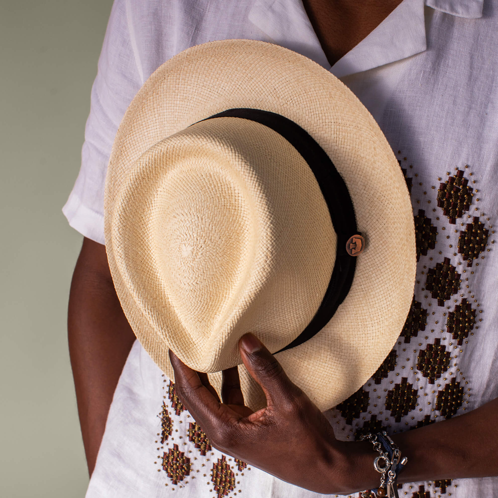 A person wearing a white patterned shirt holds the Goorin Bros. John Sr. toquilla teardrop fedora with a black band and button detail against their chest, offering stylish sun protection with 50 UPF.