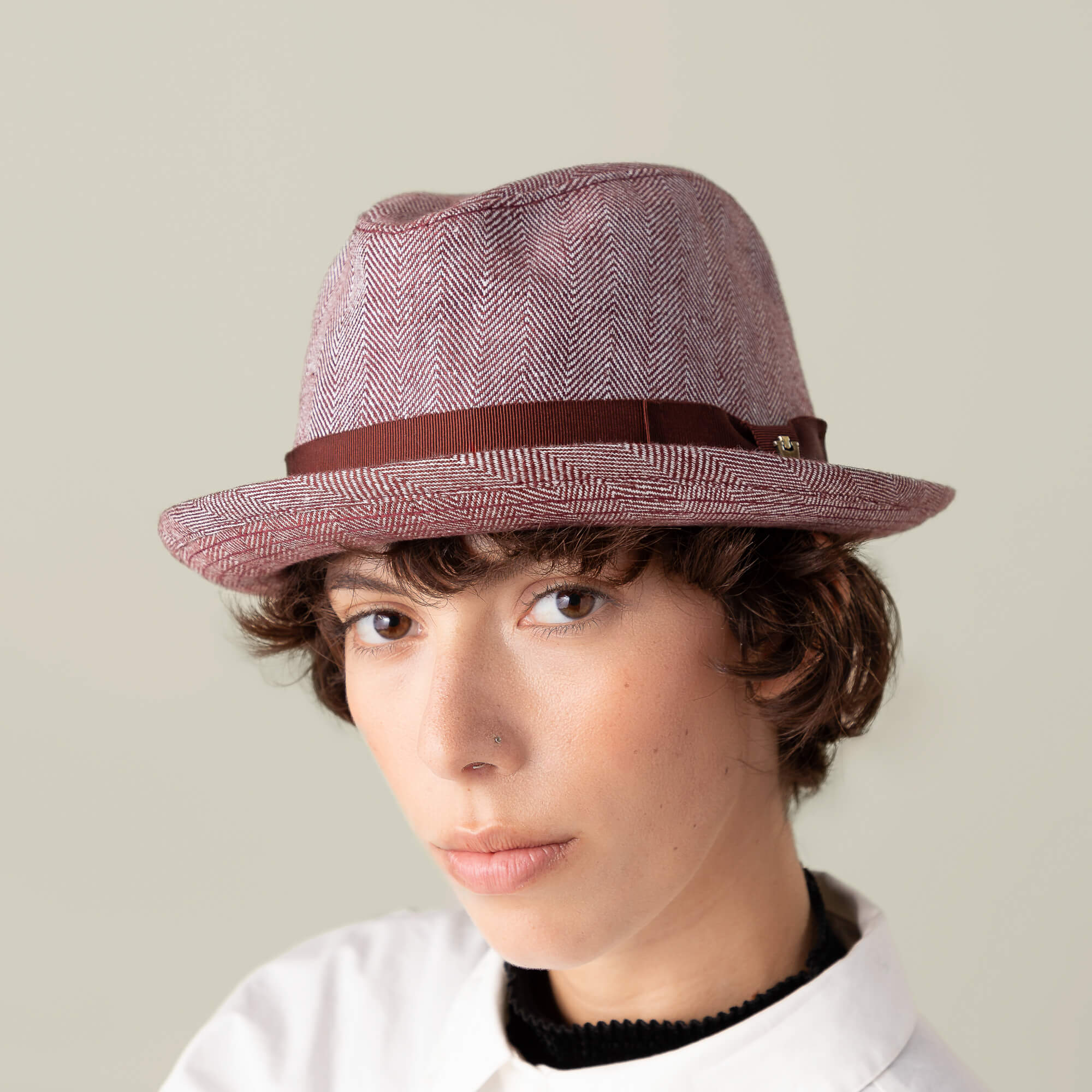 Person with short curly hair wears the Goorin Bros. Zig Zag Big Stepper, a light red structured fedora with a brown herringbone ribbon, paired with a white collared shirt and a neutral expression against a plain background.
