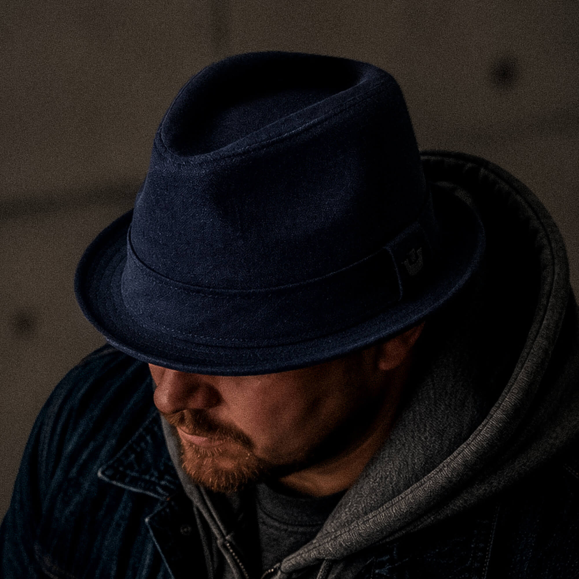 A man wearing Goorin Bros. Charlie Pants, a navy blue linen fedora, a gray hoodie, and a denim jacket looks downward in a dimly lit setting.