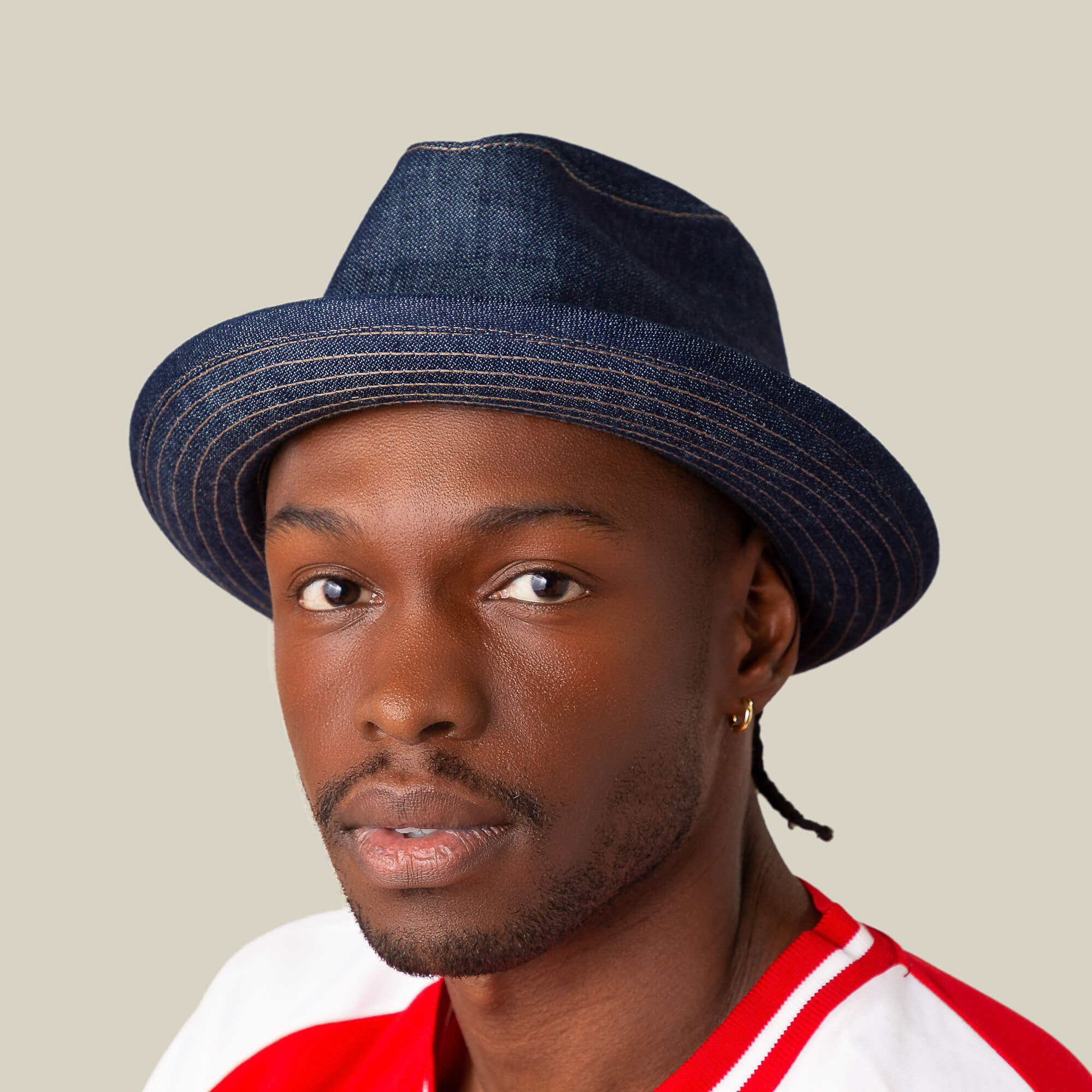 A man in a Goorin Bros. Noisy Big Stepper dark blue denim fedora with a sharp crown, small hoop earring, and red and white shirt looks directly at the camera against a plain background.