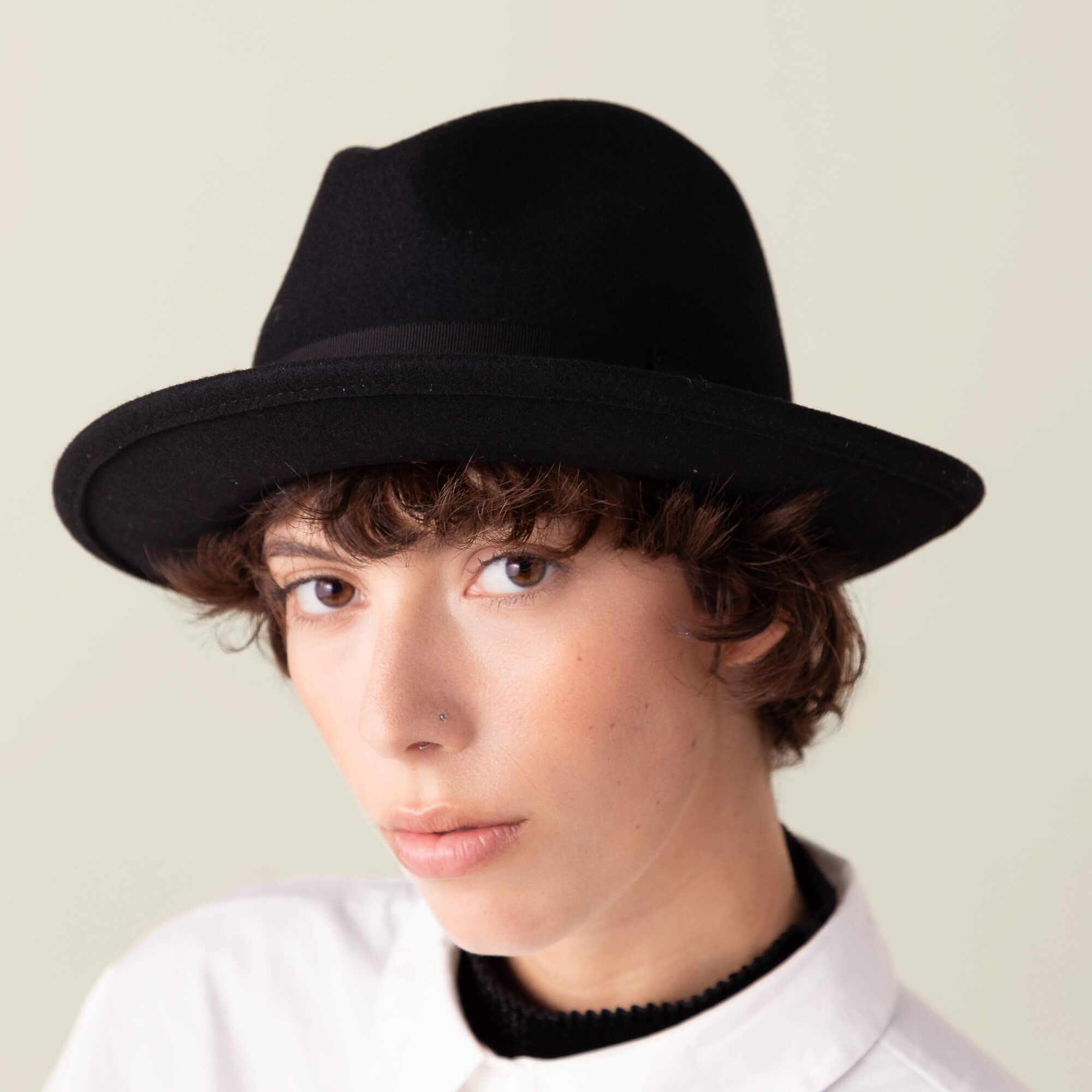Person with short curly brown hair wearing the "Dean the Butcher" snap-brim fedora by Goorin Bros. and a white collared shirt, looking at the camera with a neutral expression against a plain background.