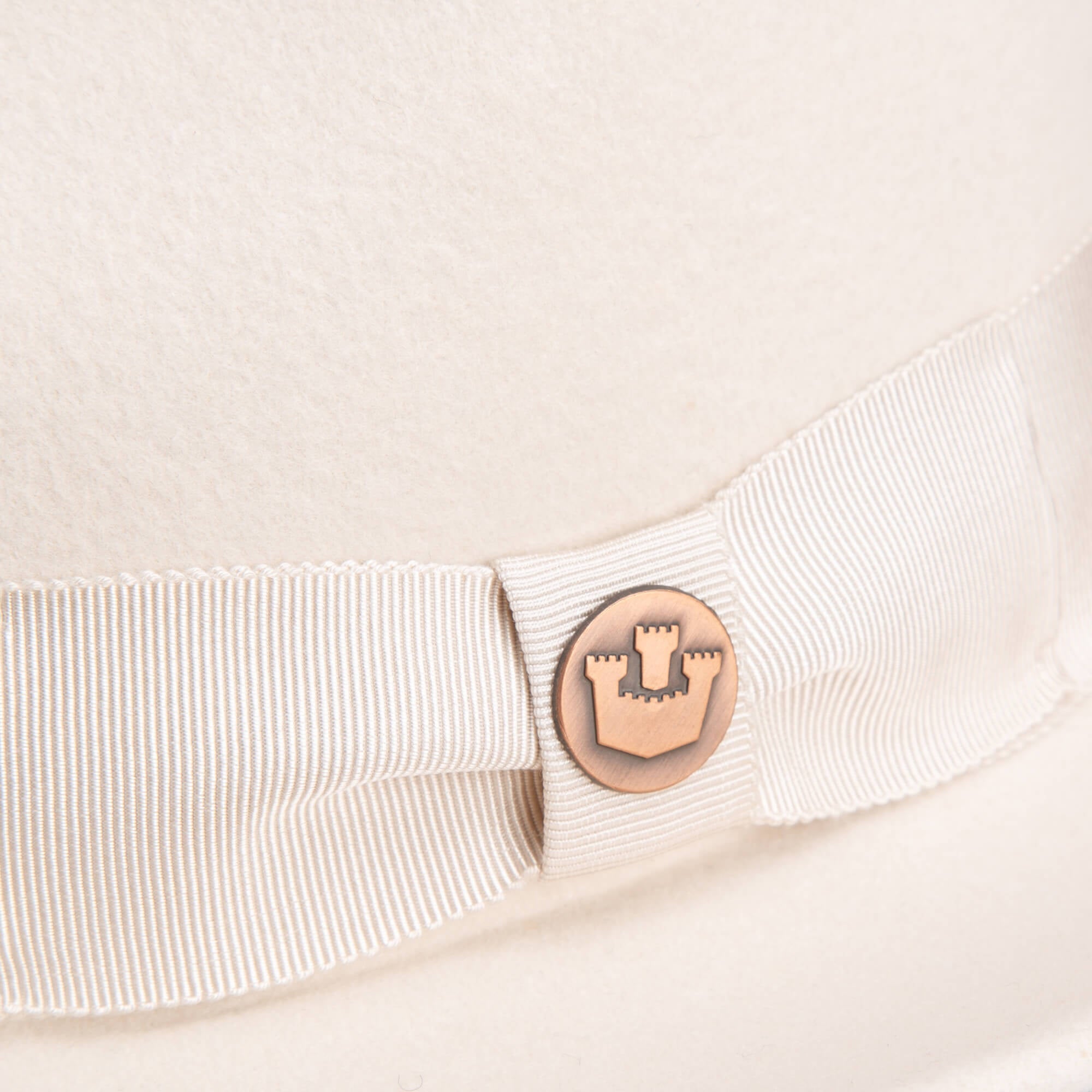 Close-up of the Goorin Bros. “Dean the Butcher” fedora in cream, featuring a matching grosgrain hatband and a round metal badge with castle emblem—an elegant hat for any season.