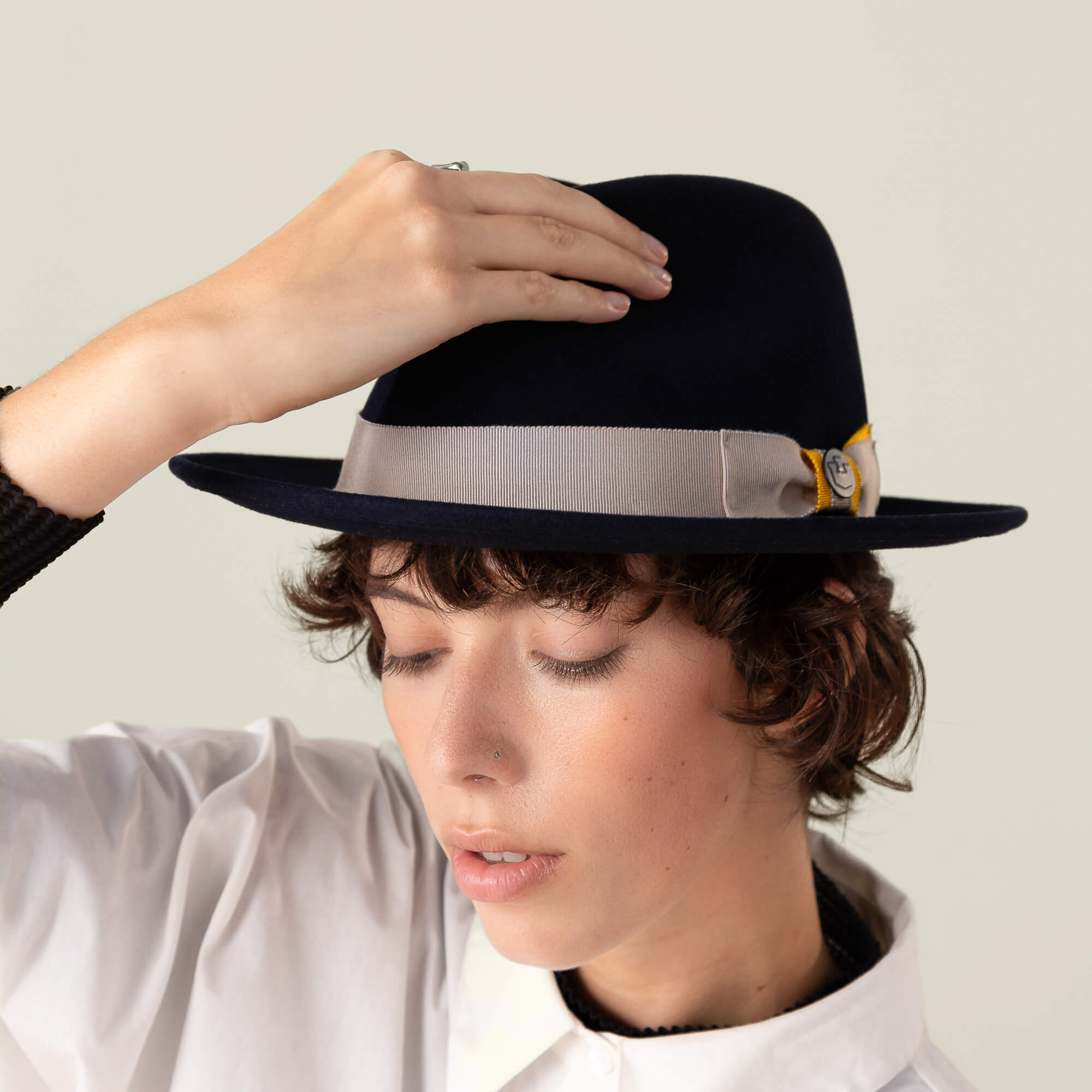 A person with short hair, dressed in a white shirt, tilts Goorin Bros.' American-made "Dean the Butcher" fedora—an all-season hat with a gray ribbon and yellow accent.
