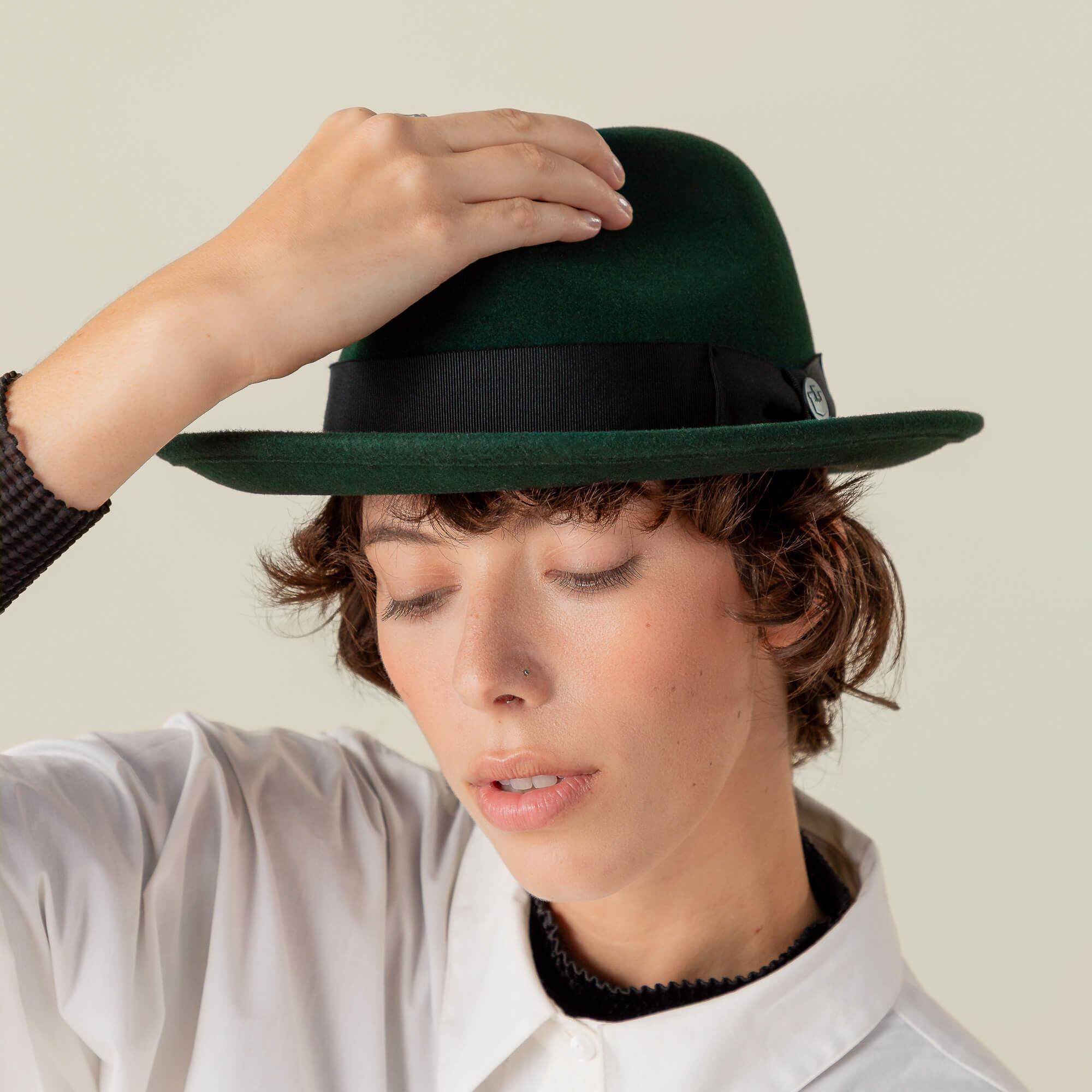 A person wears the Dean the Butcher dark green fedora by Goorin Bros. and a white collared shirt, eyes closed, with one hand touching the top of the hat.