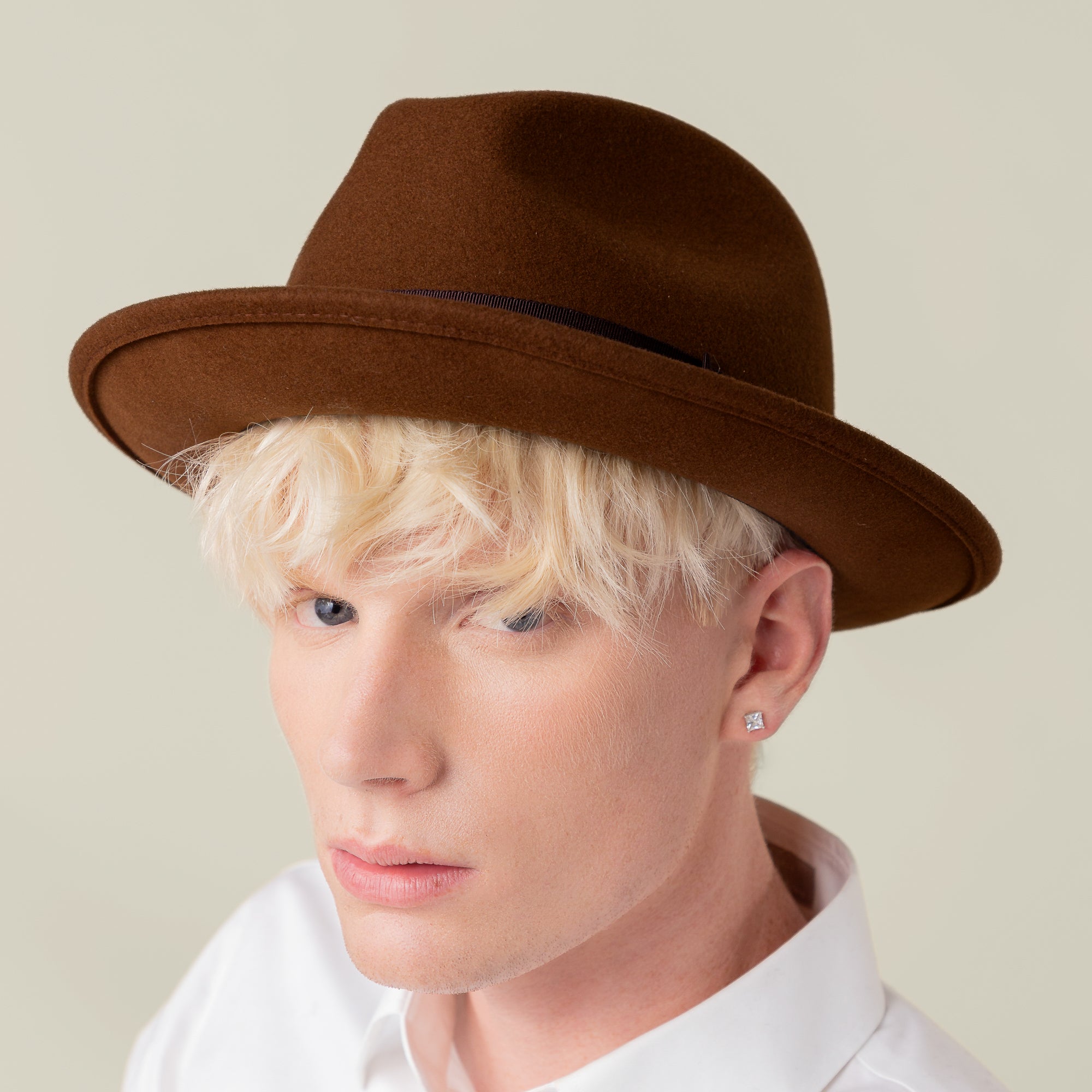A light blonde person wears the Goorin Bros. "Dean the Butcher" brown fedora, a white shirt, and a square stud earring, looking at the camera against a neutral background.