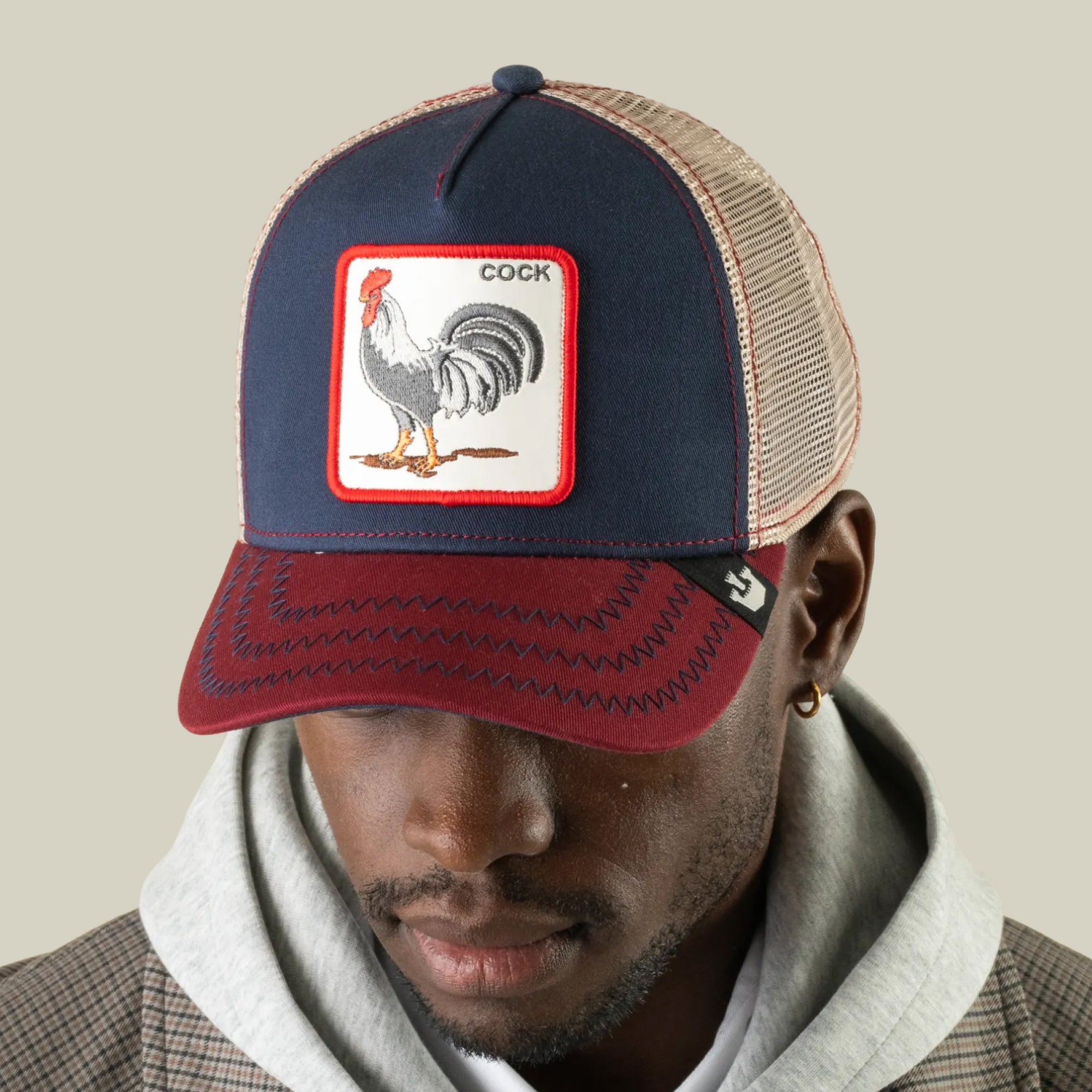 A man wearing The Farm’s “The Cock”—a navy and burgundy trucker hat featuring a rooster patch with “COCK” text, a playful symbol of sexuality and resurrection—paired with a gray hoodie and plaid jacket, looks downward against a plain background.