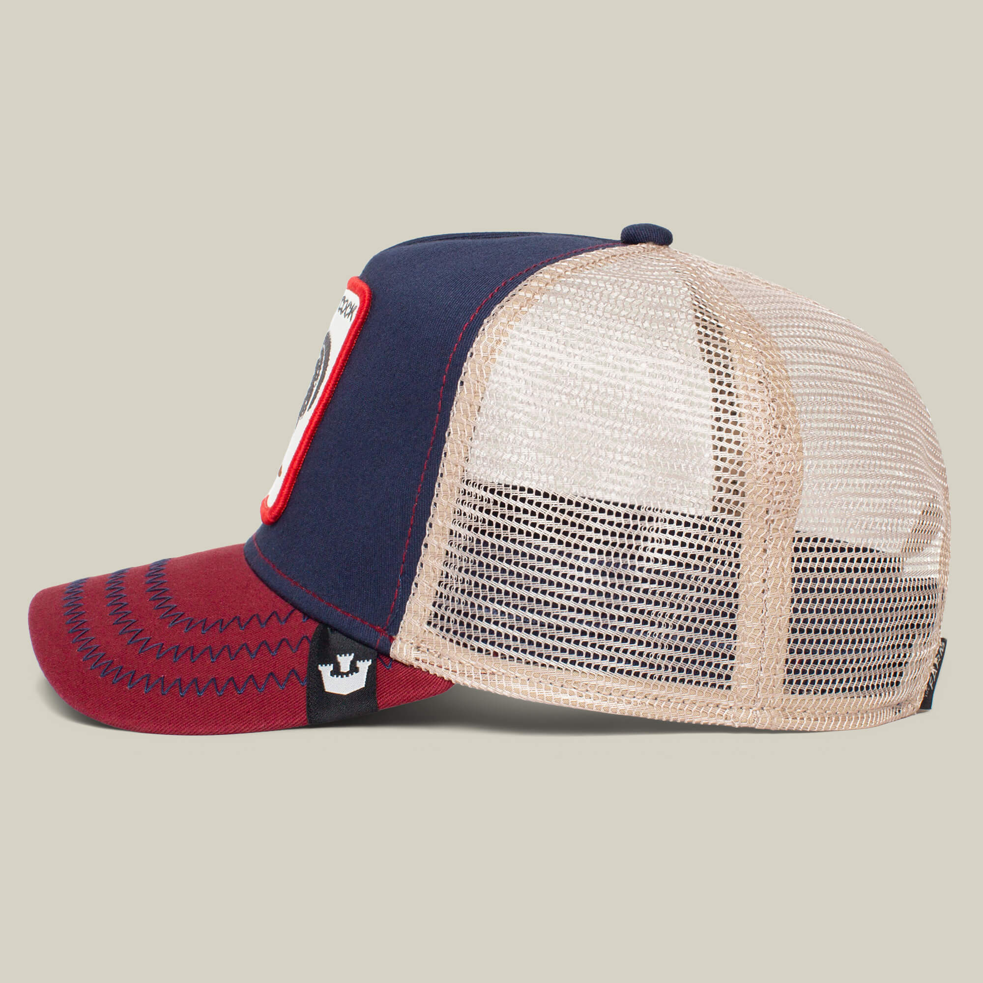 A side view of The Farm’s “The Cock” trucker hat features a navy front, burgundy brim, white rooster patch, and beige mesh back, set against a plain background.