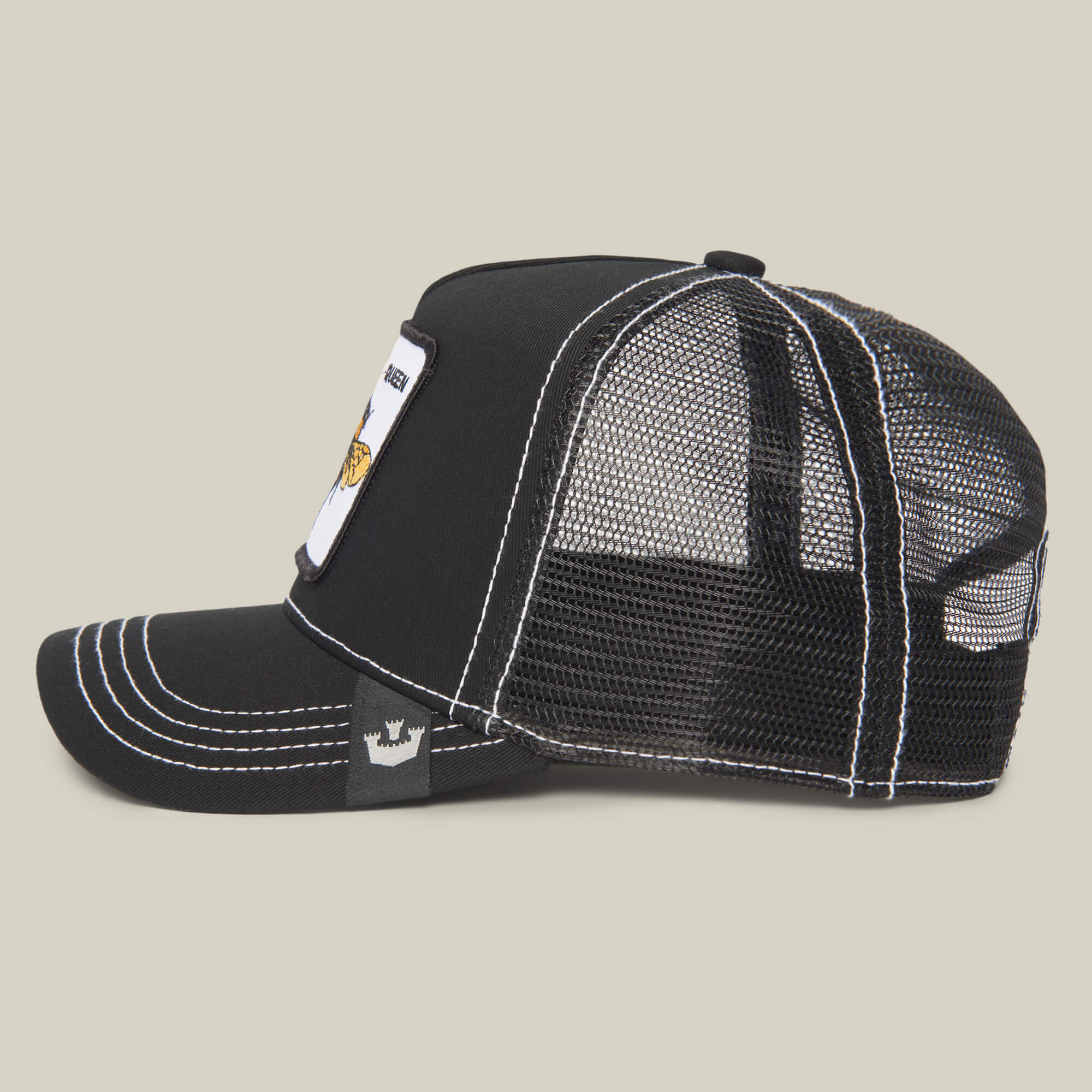 The Queen Bee by The Farm: a black mesh trucker hat with curved bill, white contrast stitching, side crown logo, and front patch—shown from the side on a beige background. Made to slay every look.