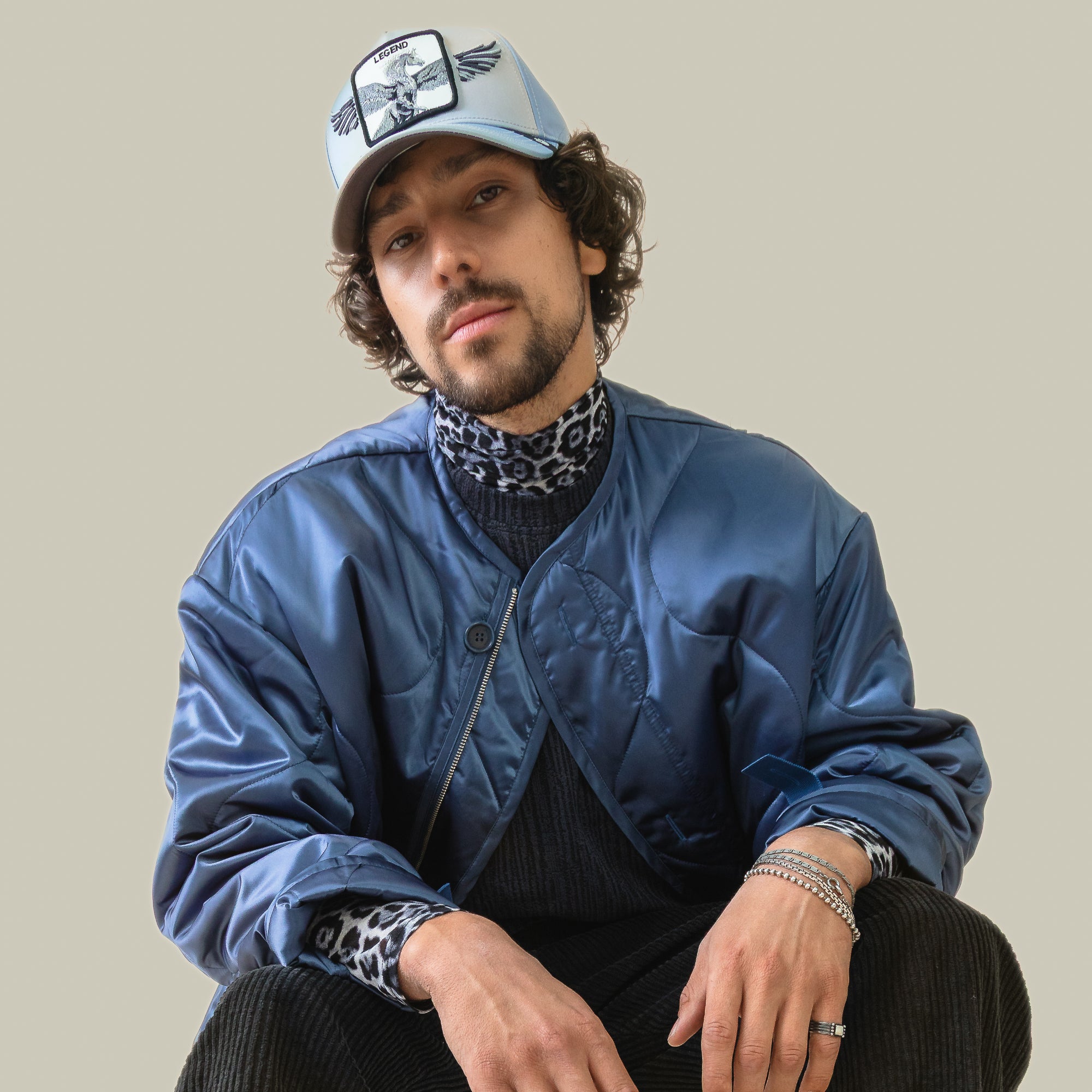 A man wears the Goorin Bros. Flare Up pale blue cap with a graphic Pegasus, paired with a light blue bomber jacket and leopard print turtleneck, seated against a plain light background.