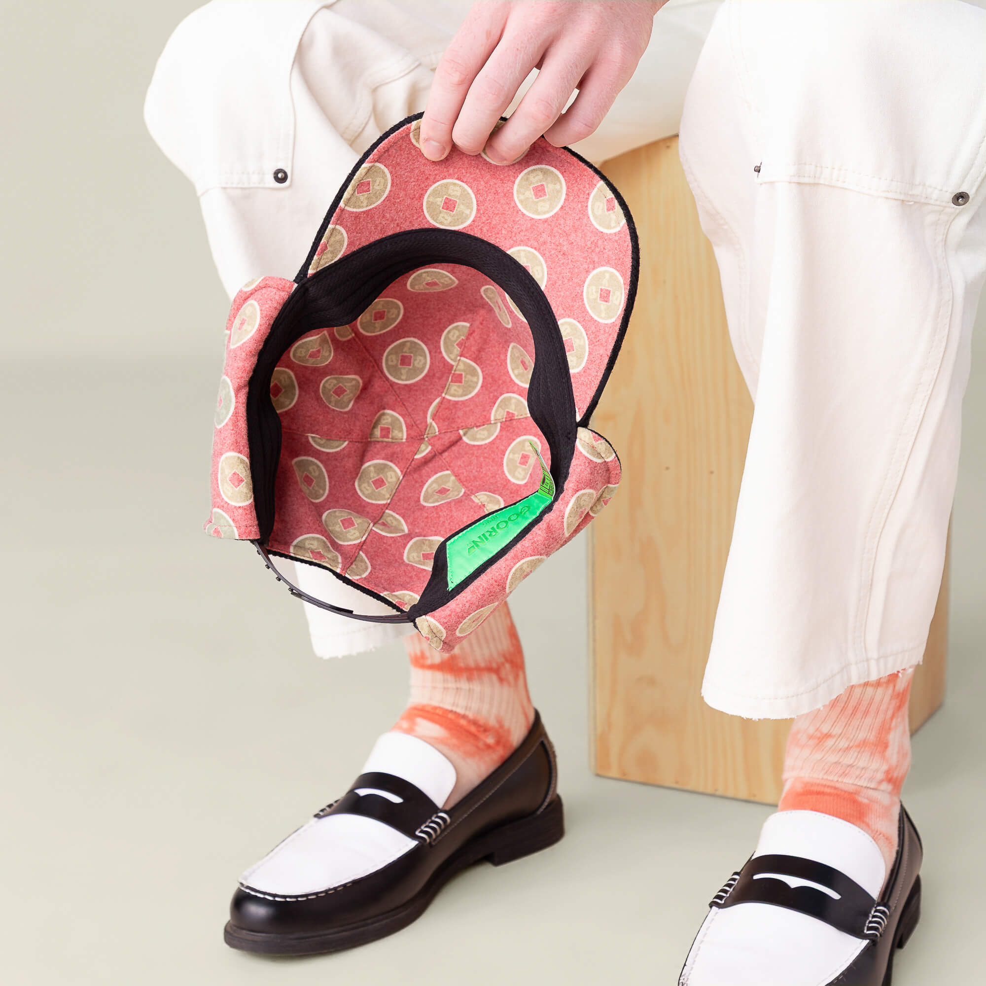 A person in white pants and orange tie-dye socks sits on a wooden block, holding the Goorin Bros. "F is for Fortune" ear-flap trucker cap with a pink and green pattern upside down.