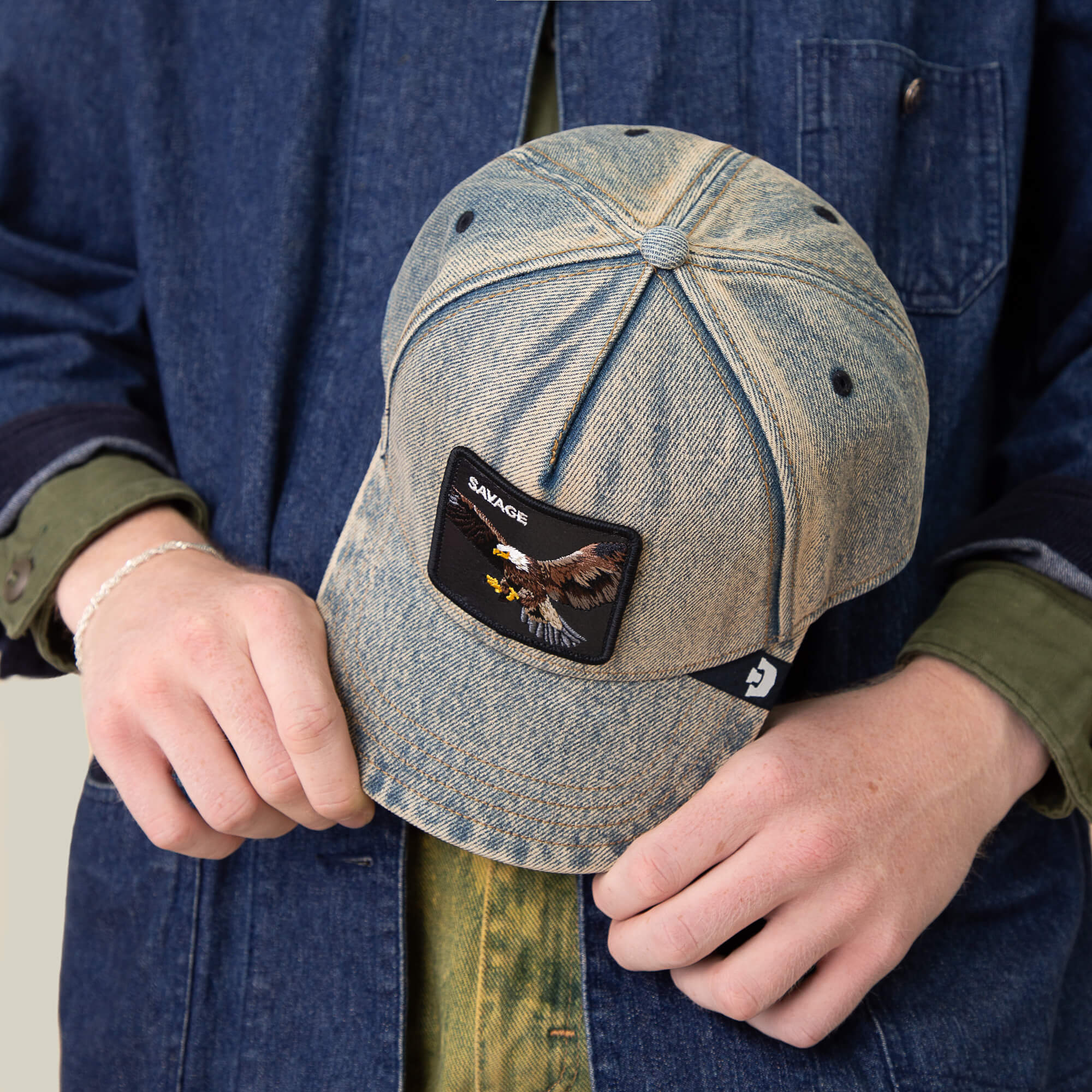 A person in a denim jacket and green shirt holds Goorin Bros.' Frequent Flyer cap, a vintage-style denim hat with an eagle patch and the word SAVAGE on the front.