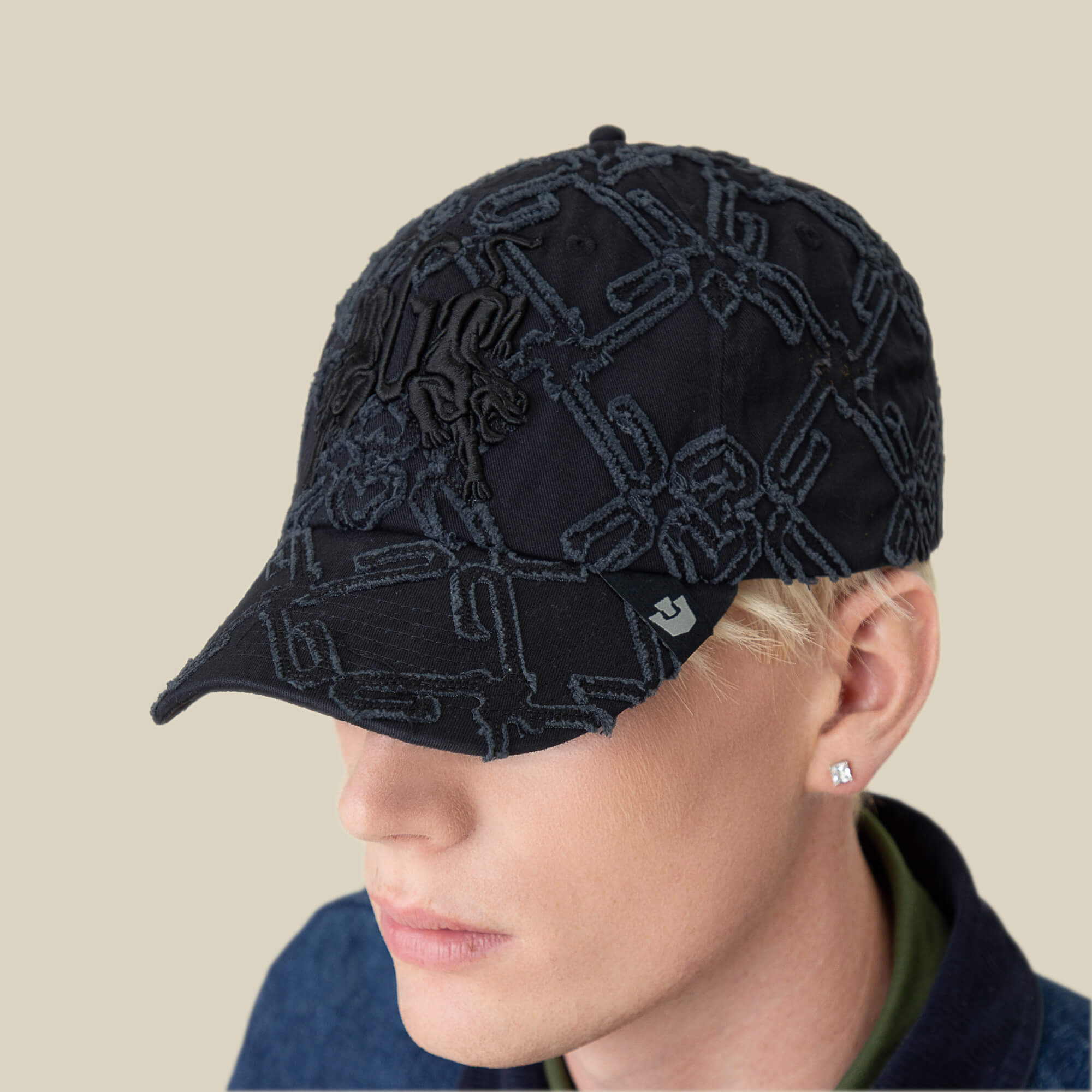 A person with short blond hair wears the Goorin Bros. Future Baby Daddy Papa Cap with all-over embroidery and a stud earring, looking partially downward against a beige background.