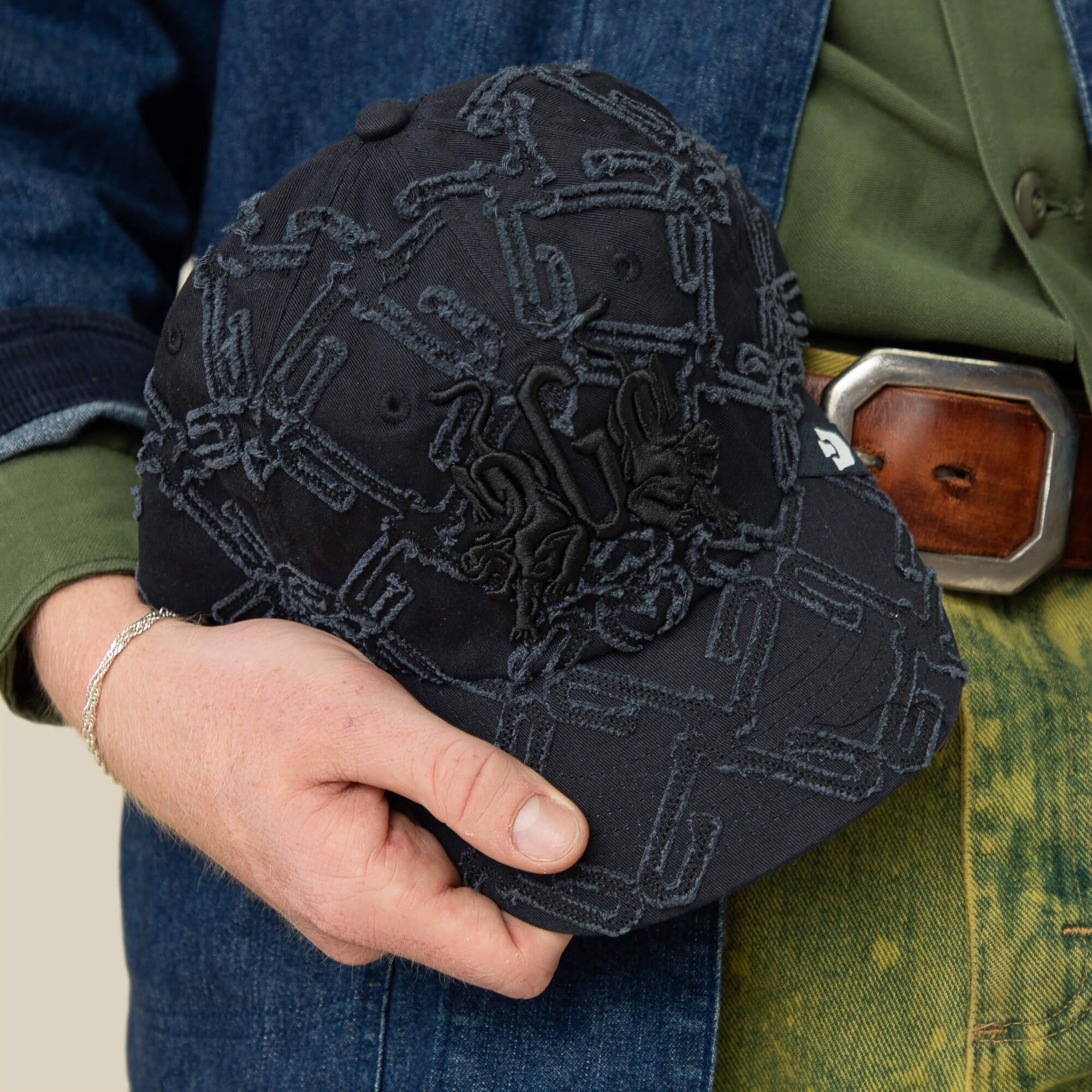 A person holds a black Goorin Bros. Future Baby Daddy Papa Cap with all-over embroidery and textured abstract patterns, paired with a green shirt, denim jacket, and a brown belt with a rectangular buckle.
