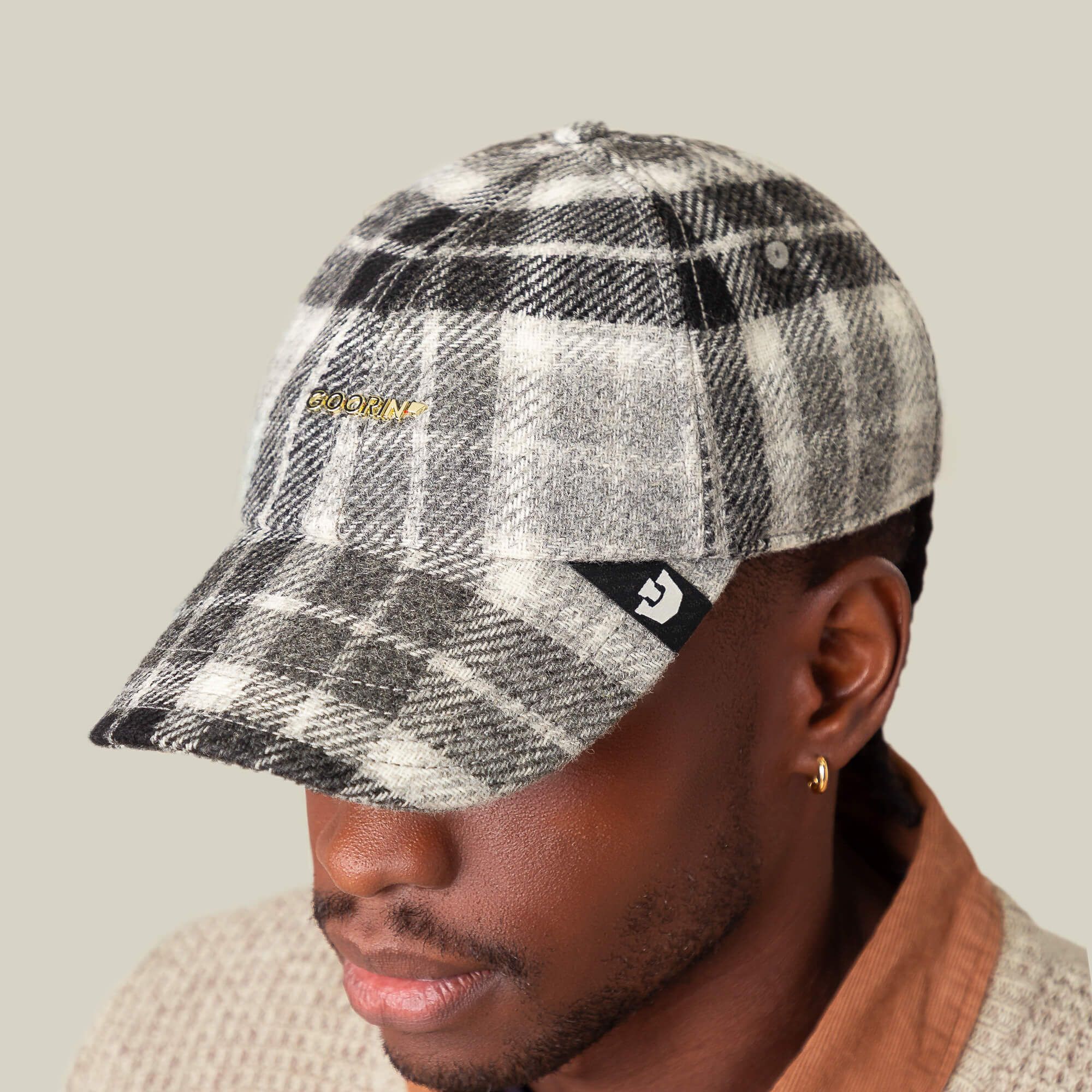 A person wearing the Goorin Bros. Bespoke Wordmark cap—a black and white plaid wool blend hat featuring a small logo patch on the side and gold embroidered text on the front.