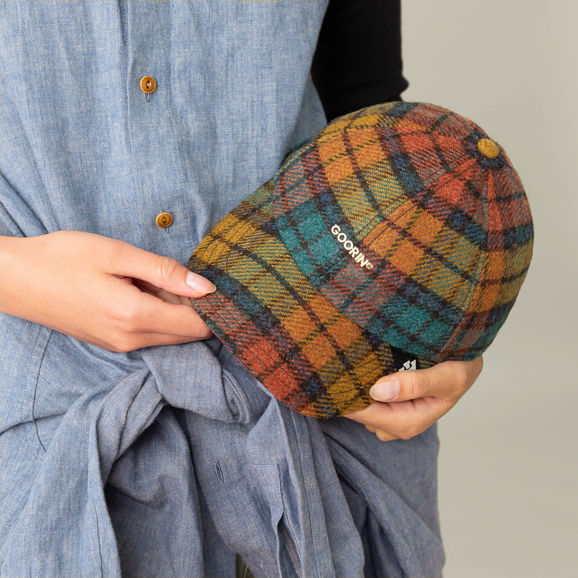 A person in a blue button-down dress holds the Goorin Bros. Bespoke Wordmark plaid wool blend cap, featuring "GOORIN" embroidered on the side.