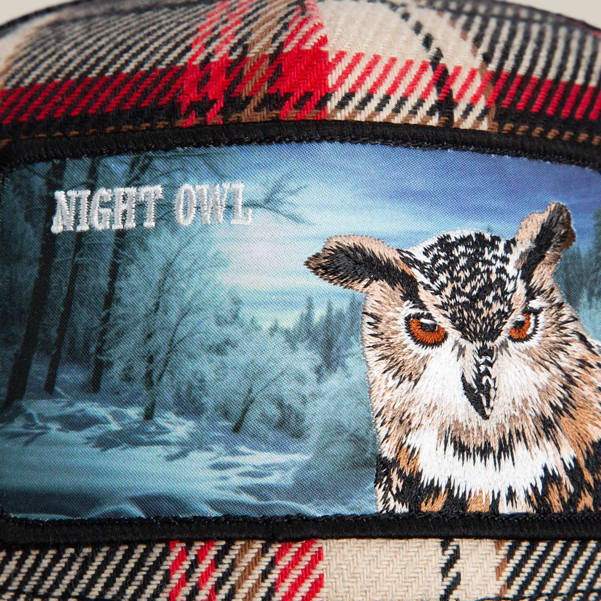 The Goorin Bros. Cabin Fever flannel plaid fabric showcases a patch with an embroidered owl, a wintry forest backdrop, and "NIGHT OWL" in white text, capturing rugged comfort and woodland charm.