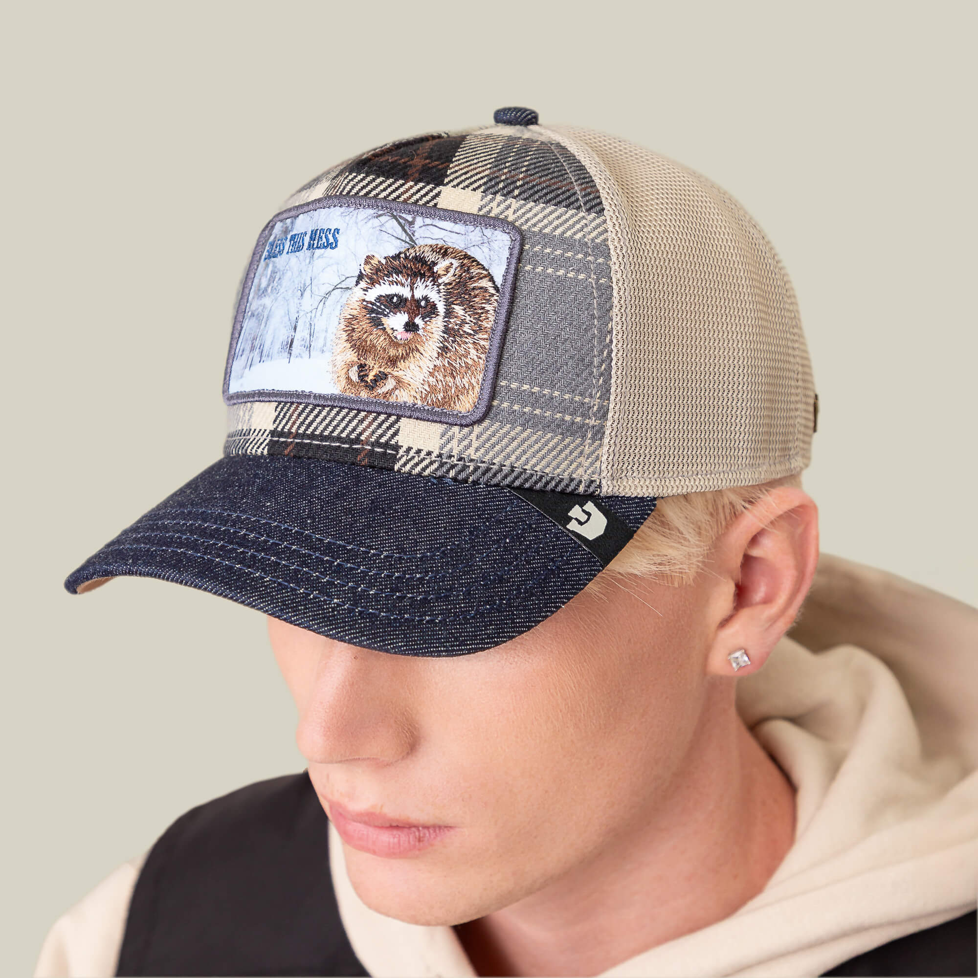 A person wearing the Goorin Bros. "Free Spirited" mesh baseball cap, featuring a plaid crown, denim brim, and raccoon patch, looks downward.