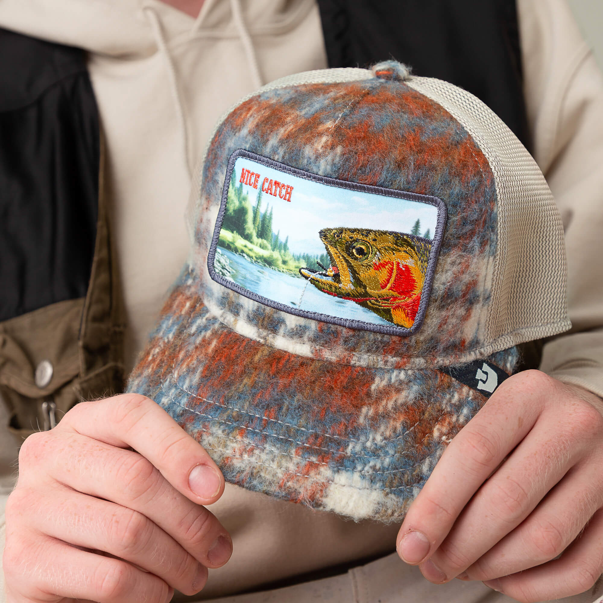 Someone holds the Goorin Bros. "Down by the River" mesh trucker hat, featuring a fish patch, against a nature backdrop—perfect for your next fishing trip or pairing with a cozy flannel outdoors.