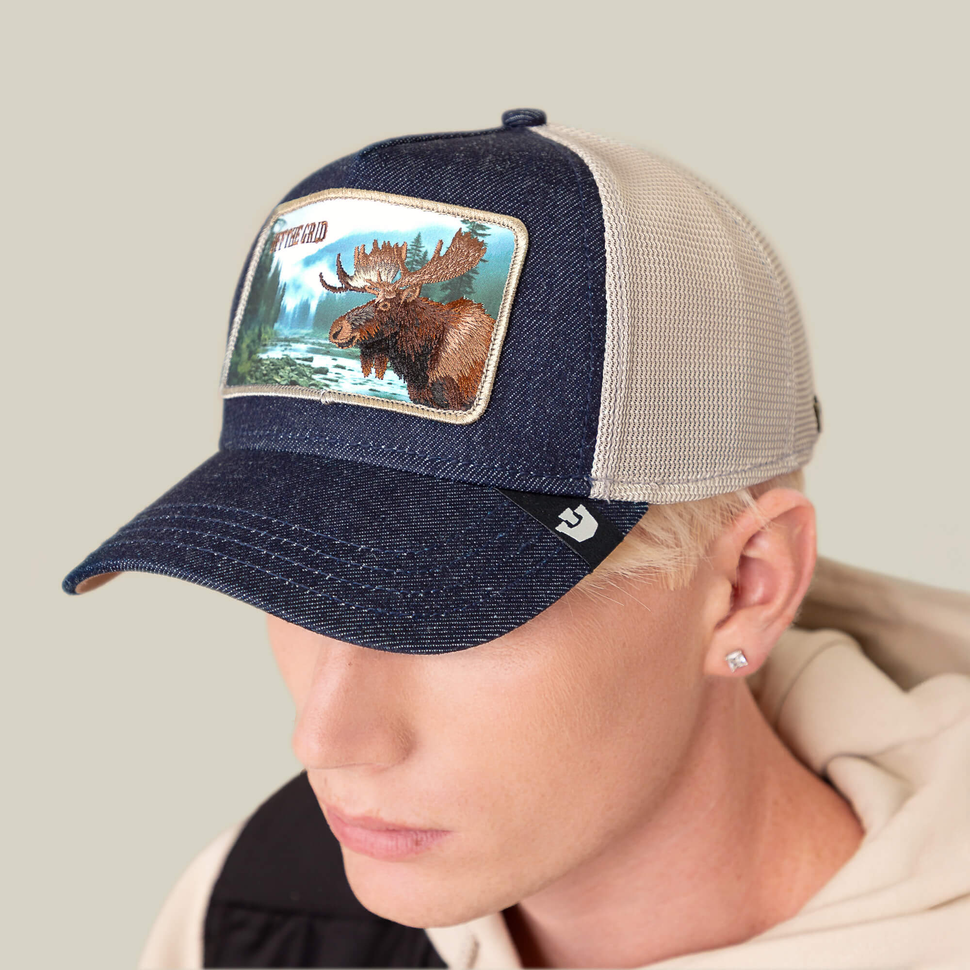 A person wearing the Goorin Bros. "Staycation" blue and beige mesh trucker hat, featuring a moose patch with a wilderness scene and mountains on the front.