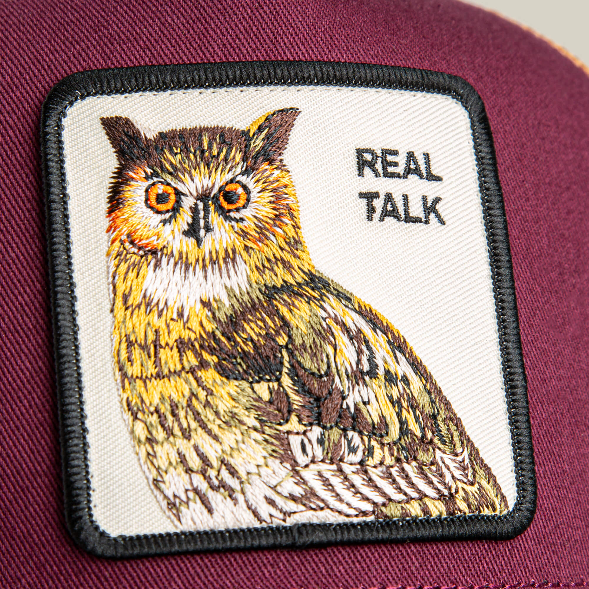 A close-up of the Goorin Bros. Tricolor Owl patch cap, showing an illustrated owl and the words REAL TALK against a rich fabric background.