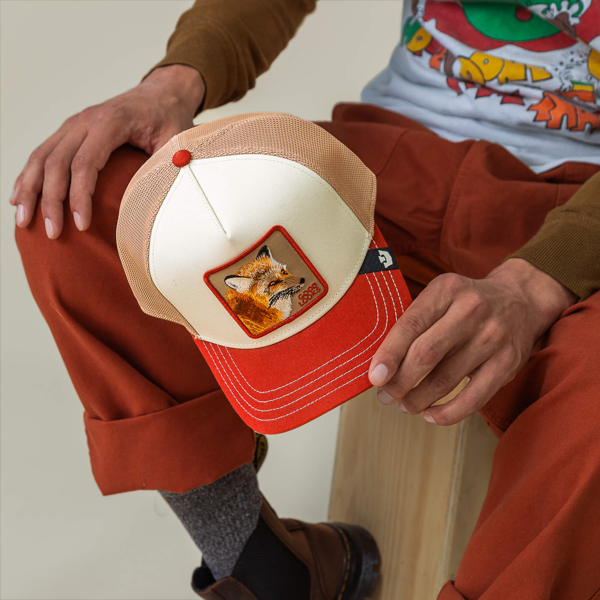 A person sits on a wooden block, holding the Goorin Bros. Tricolor Fox trucker hat featuring a fox patch. They wear rust-colored pants, brown shoes, and a graphic t-shirt.