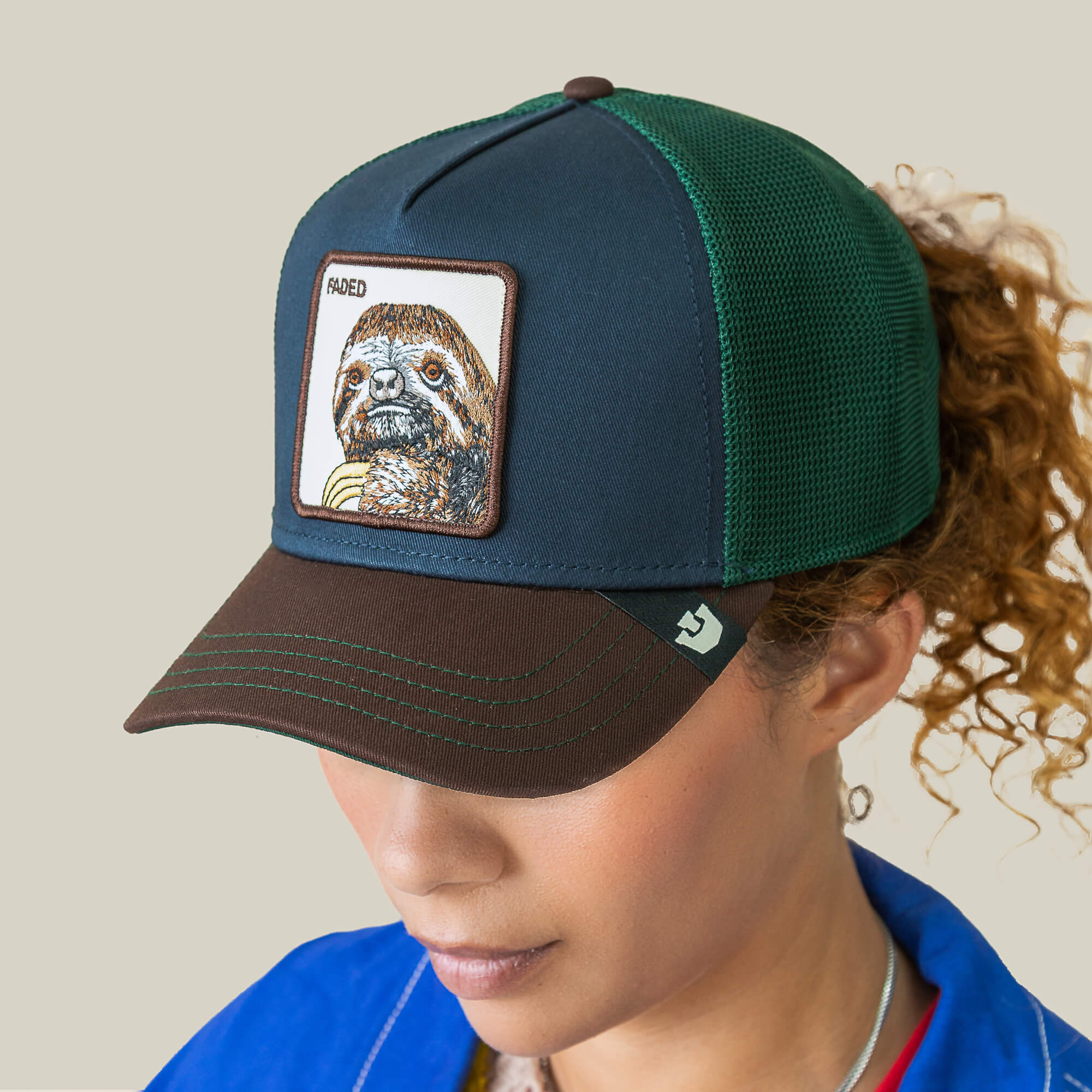 A person wearing the Goorin Bros. Tricolor Sloth hat—a green and brown cap with mesh panels and a sloth patch labeled SLOWED—a fun, playful fashion choice.
