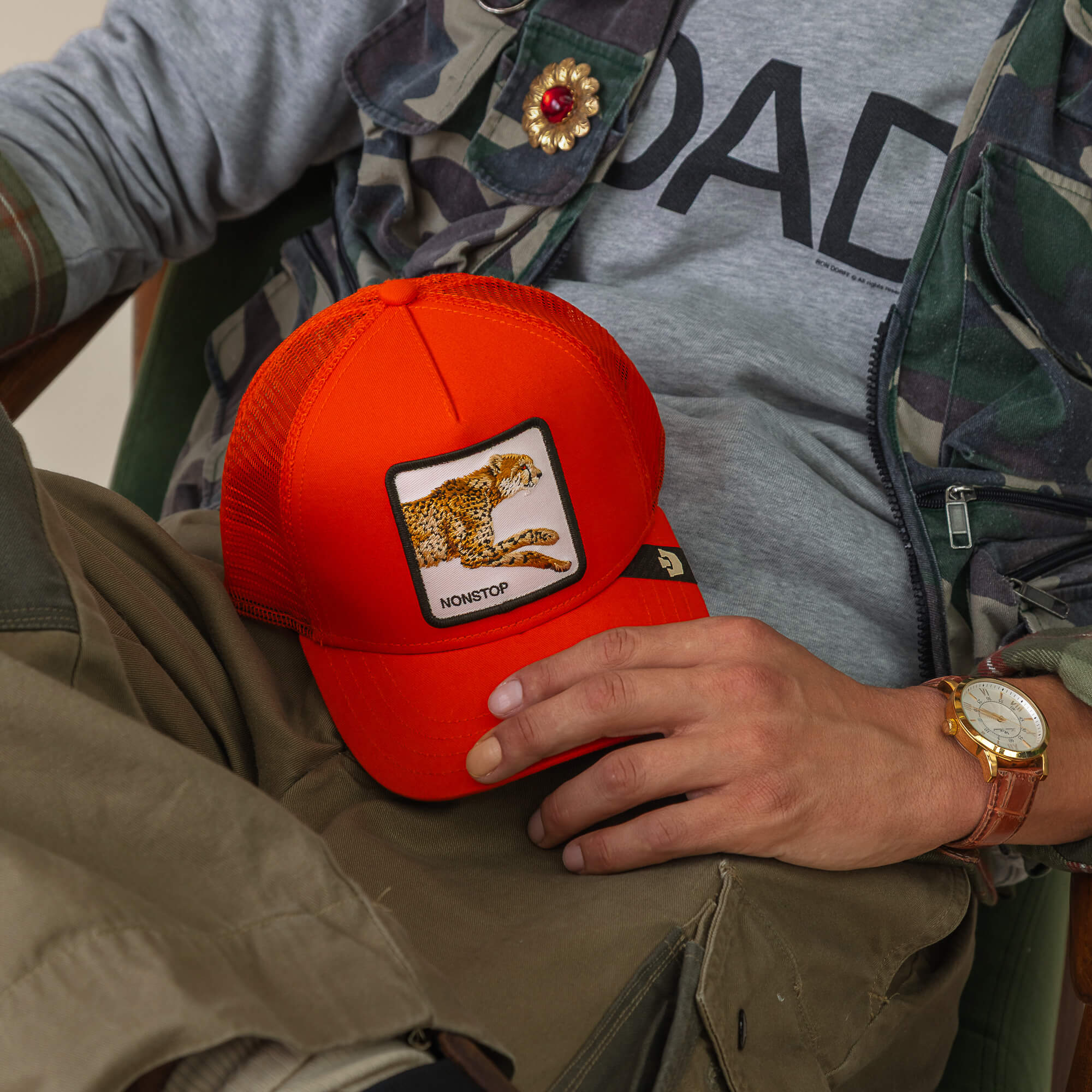 A person holds Goorin Bros.’ The Nonstop Cheetah, a vivid orange trucker cap with a leopard patch and "NONSTOP," styled with a camo jacket, gray shirt, and brown watch.