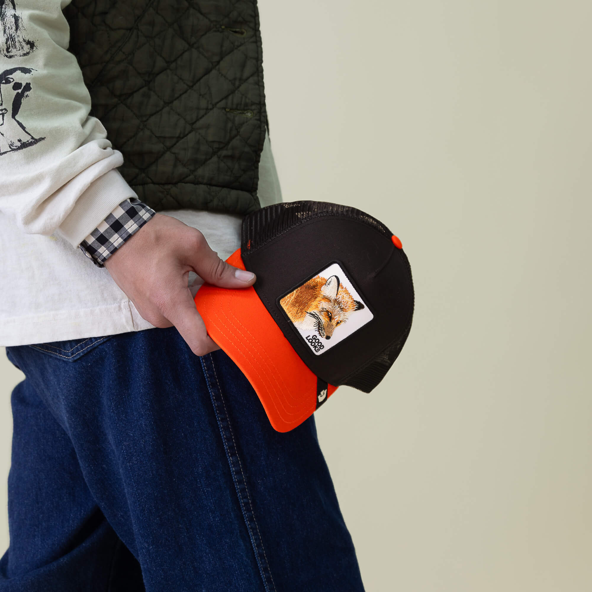 Person holding the Goorin Bros. Good Looks Fox trucker hat with a bold fox patch, wearing a white shirt, green vest, and blue jeans for a farmyard chic vibe.