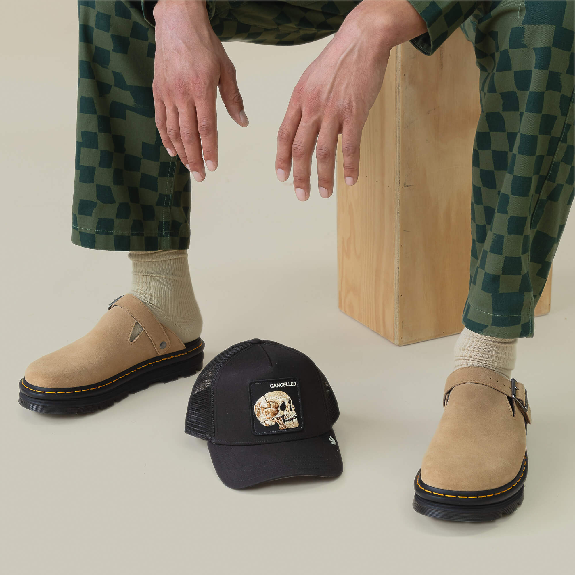 A person in checkered green pants, beige socks, and tan clogs sits on a wooden block. Nearby, the Goorin Bros. "The Cancelled Skull" black trucker hat with skull graphic and "speak your mind" message rests on the floor.