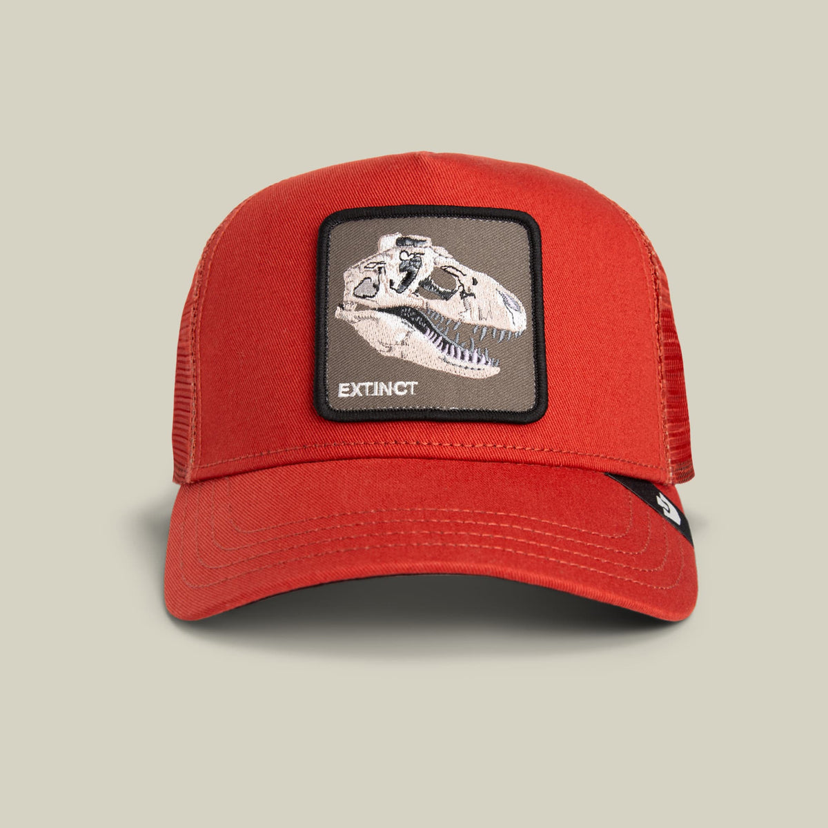 The Extinct Skull by Goorin Bros is a red trucker hat with mesh panels featuring a embroidered dinosaur skull patch on the front and the word Extinct, displayed against a neutral beige background.