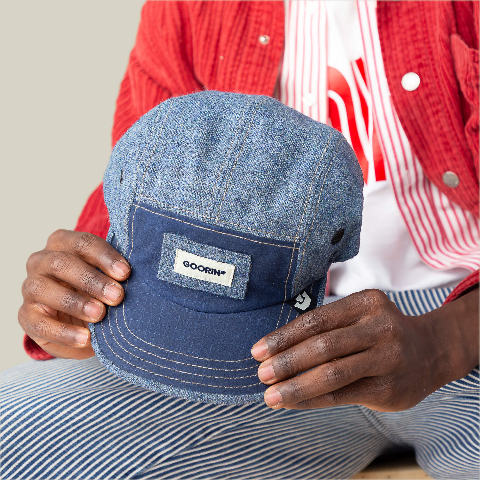 A person in a red jacket and striped pants holds the Billed Demand cap by Goorin Bros., featuring a blue denim texture and a signature Goorin patch on the front.