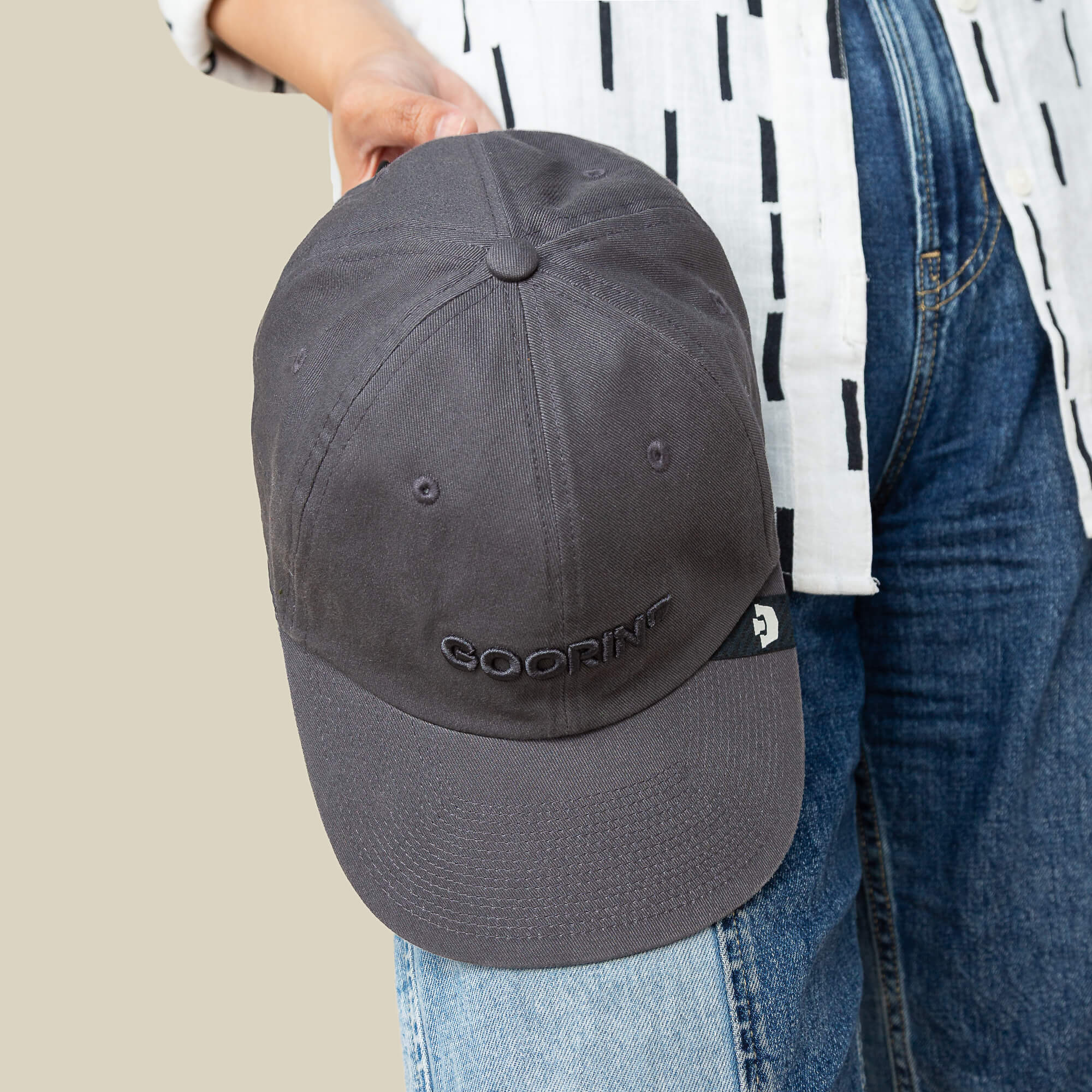 A person holds a Goorin Bros. Papa Core dark gray cap with a soft crown and curved brim, featuring "GOORIN" embroidered on the front, paired with a white patterned shirt and blue jeans.