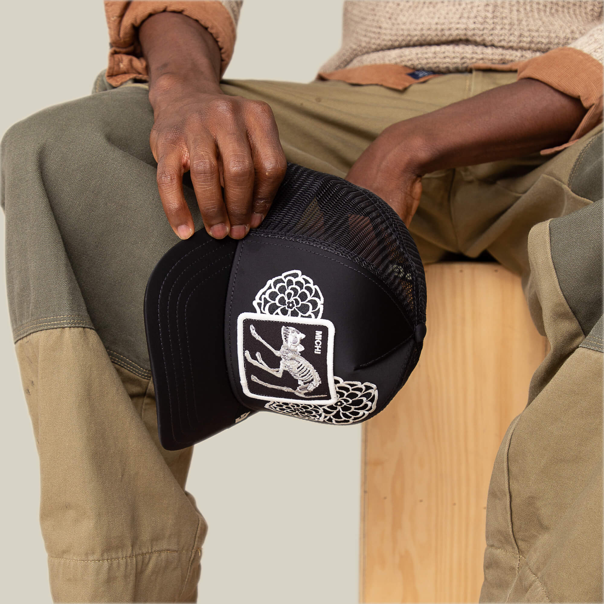 A person seated on a wooden block holds the Goorin Bros. Dia De Michi black mesh trucker hat, featuring a white crown and hand graphic patch inspired by Michi's oversized florals style.