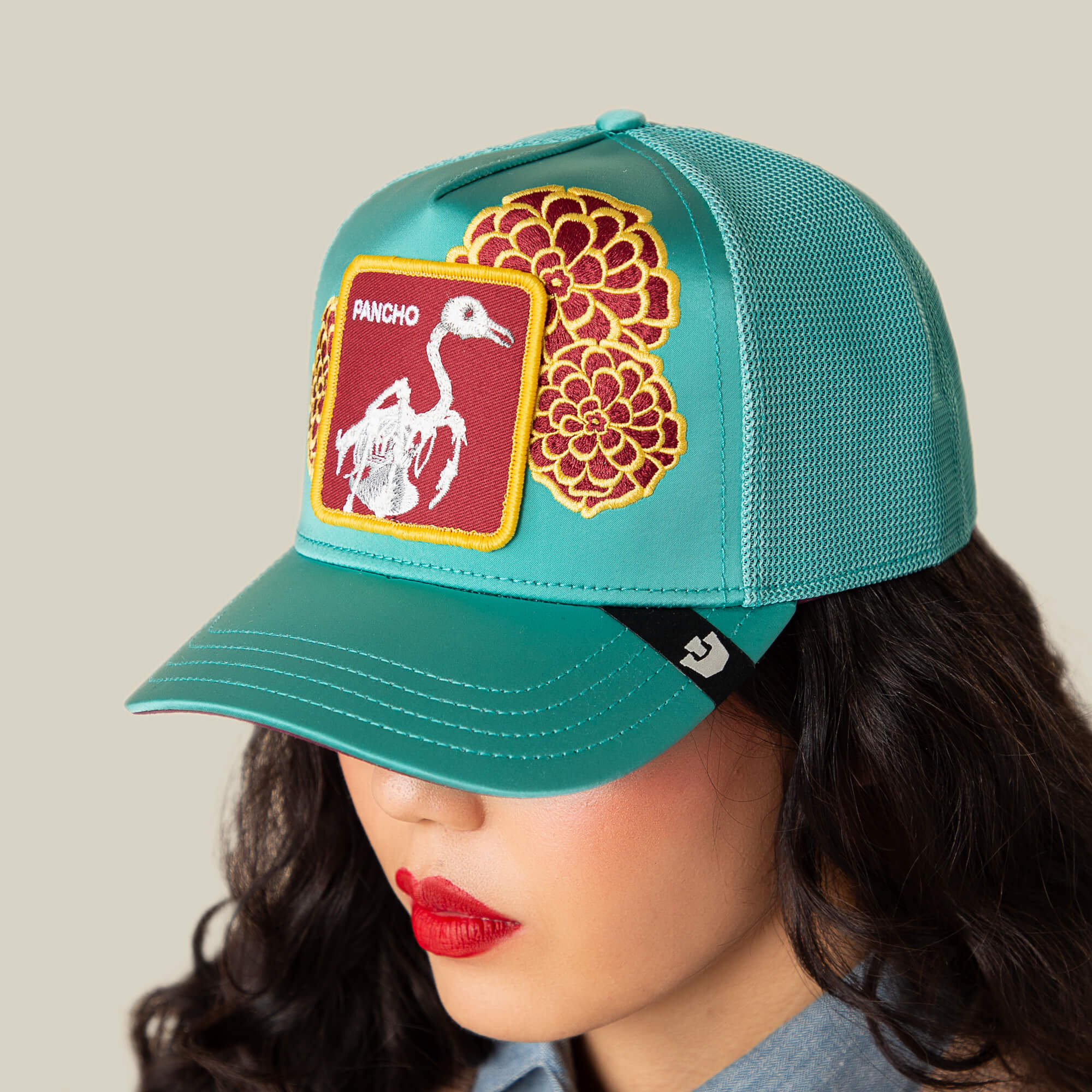 Wearing the Goorin Bros. "Dia De Pancho" teal mesh cap with floral embroidery and a skeleton bird patch, this person channels a rebel spirit, their face partly hidden beneath the hat.