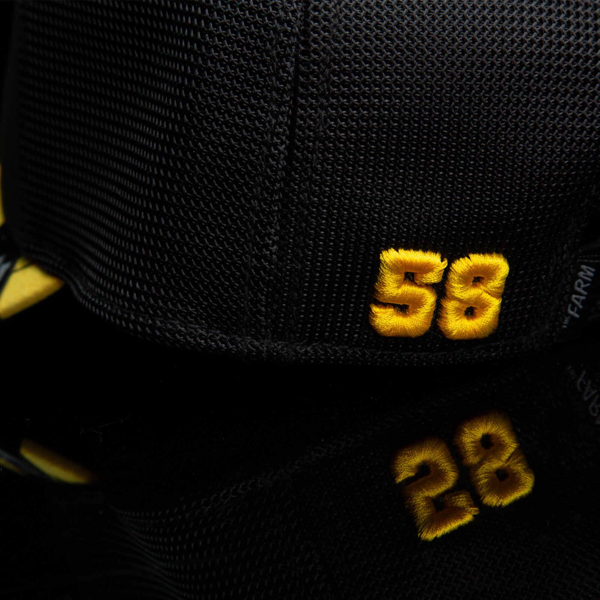Close-up of the Goorin Bros. Suede Tanger 58 black mesh cap with yellow embroidery, reflected on a glossy black surface.