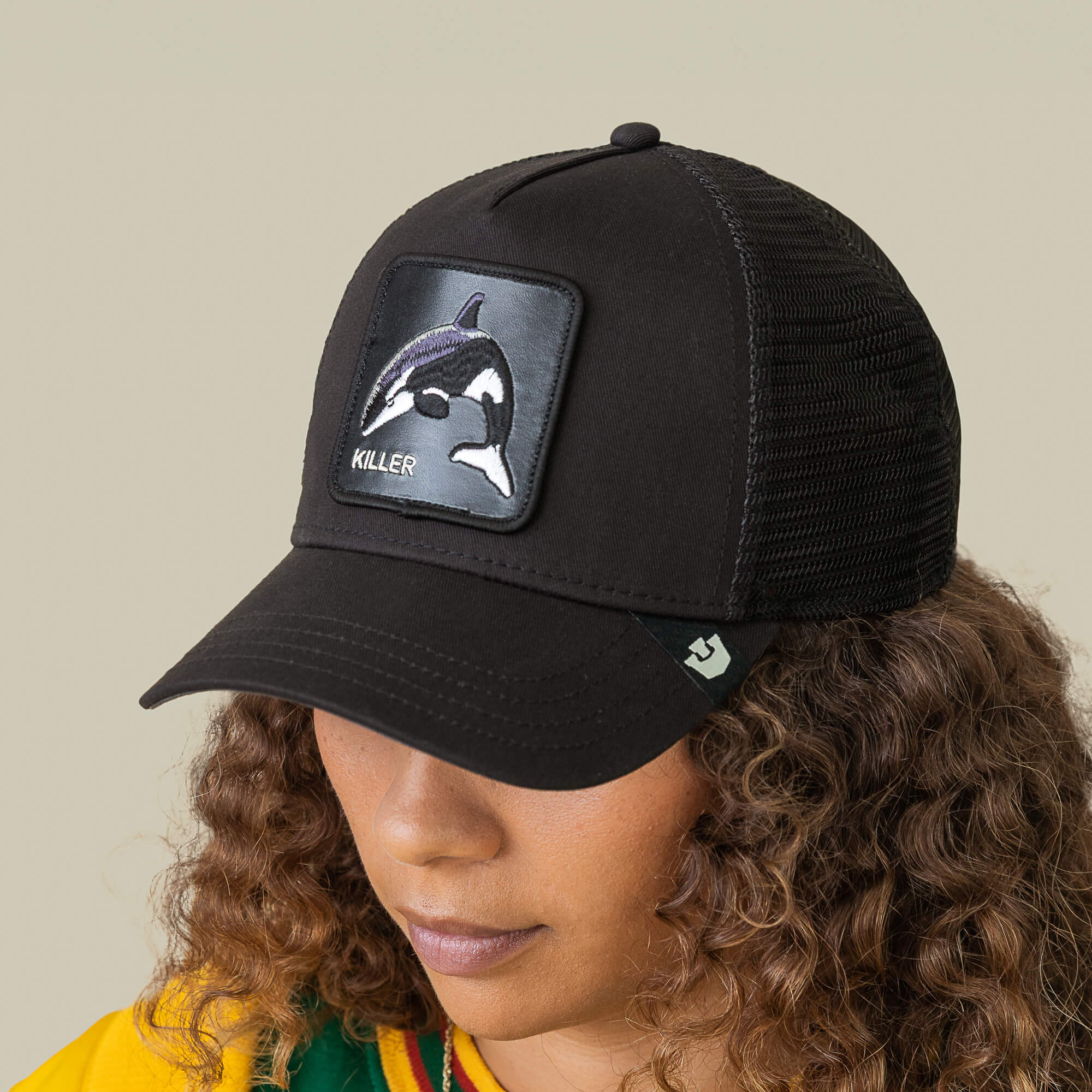 A person with curly hair looks downward wearing the Goorin Bros. Killer Trucker cap, a black and white hat featuring an embroidered orca patch with "KILLER" on the front.