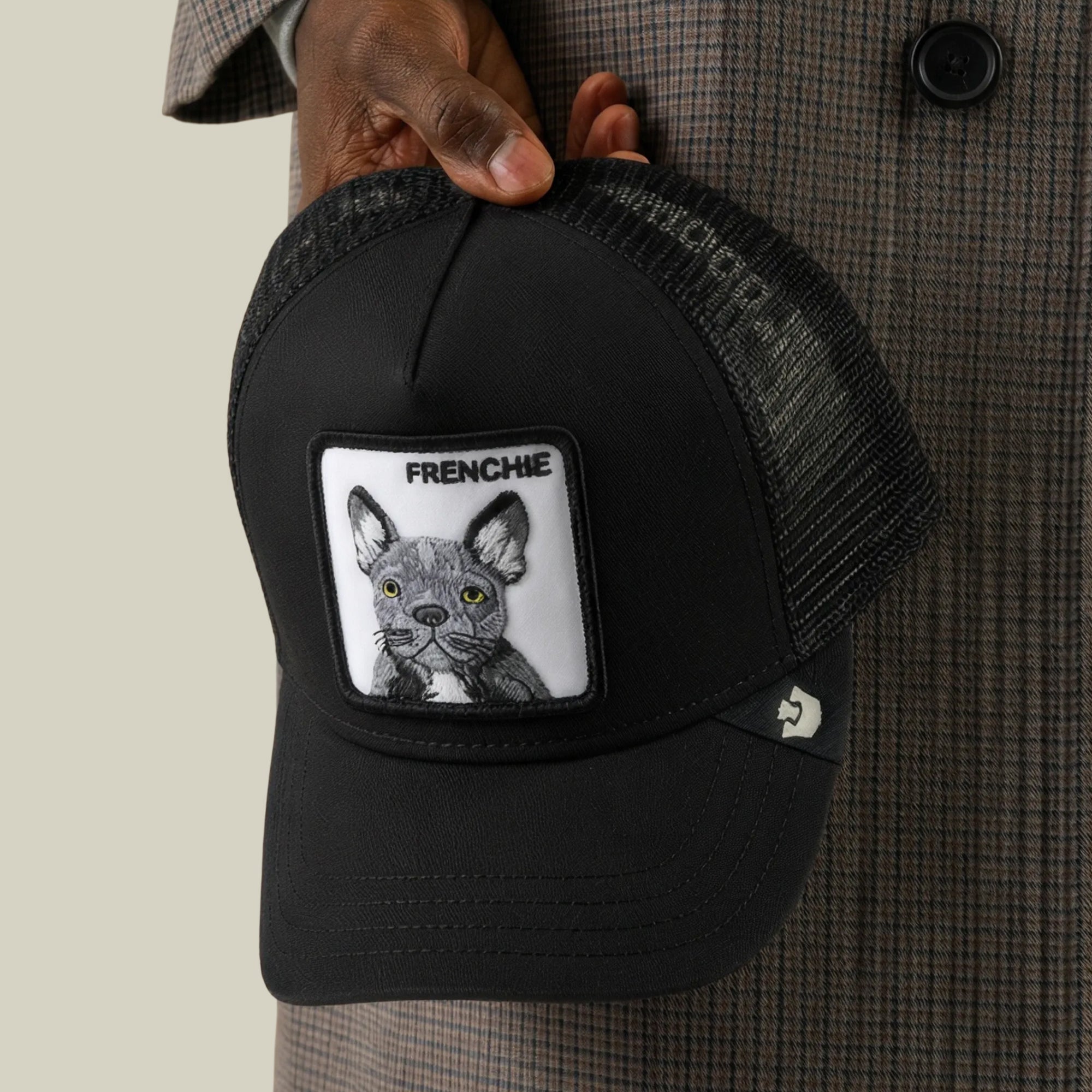 Someone wearing a plaid jacket holds the Frenchie Trucker hat by Goorin Bros., a black mesh trucker cap featuring a patch with an illustration of a French Bulldog and the word “FRENCHIE” above it.