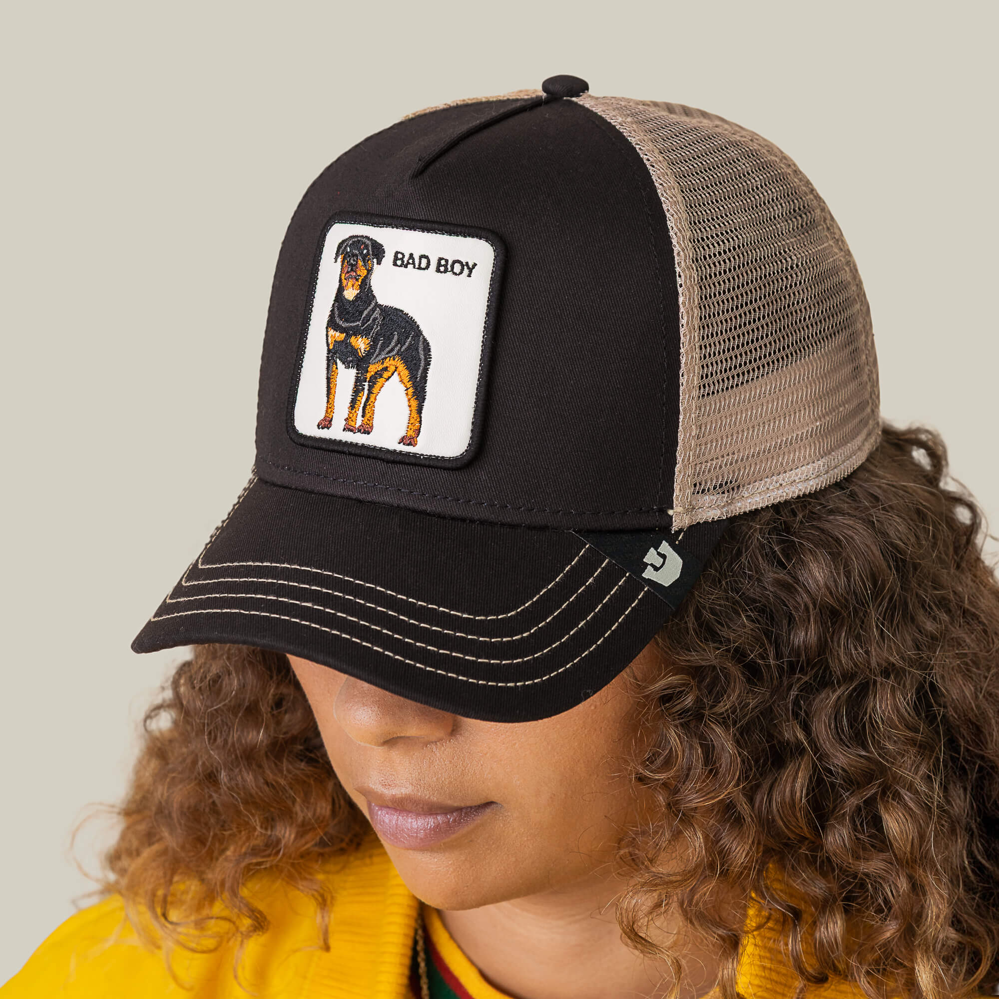 A person with curly hair looks down while wearing "The Bad Boy" mesh hat by Goorin Bros., featuring a black and beige design with a Rottweiler patch and BAD BOY text.
