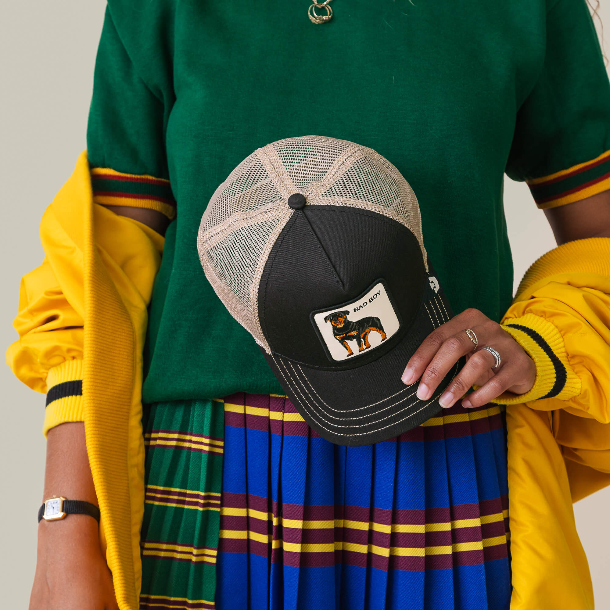 Person holding "The Bad Boy" cap by Goorin Bros.—a black and beige hat with a dog patch—paired with a green top, yellow jacket, and pleated blue striped skirt.