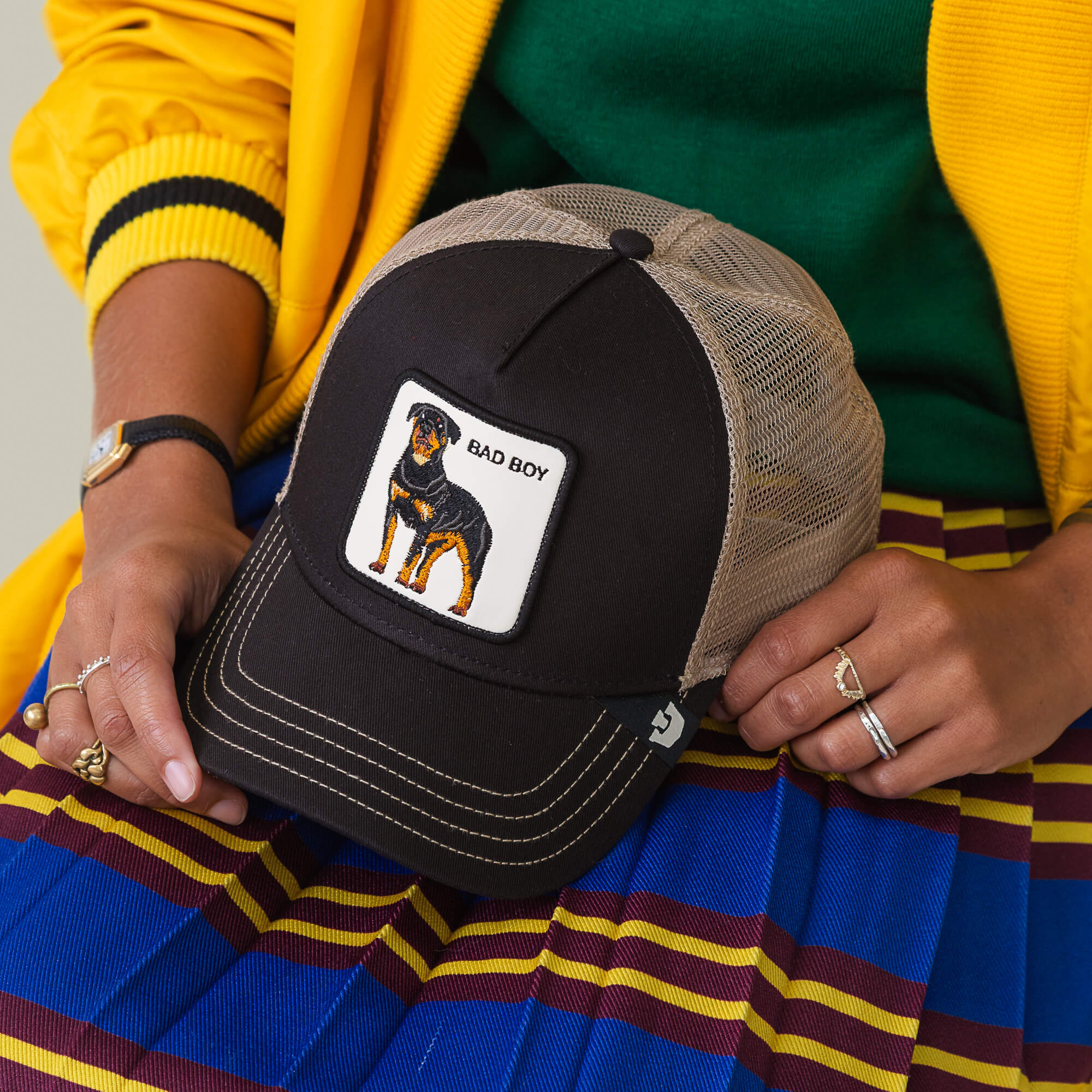 A person holds The Bad Boy hat by Goorin Bros.—a stylish black and beige mesh cap with a Rottweiler patch labeled "BAD BOY"—while wearing a yellow jacket and a blue skirt with maroon and yellow stripes.