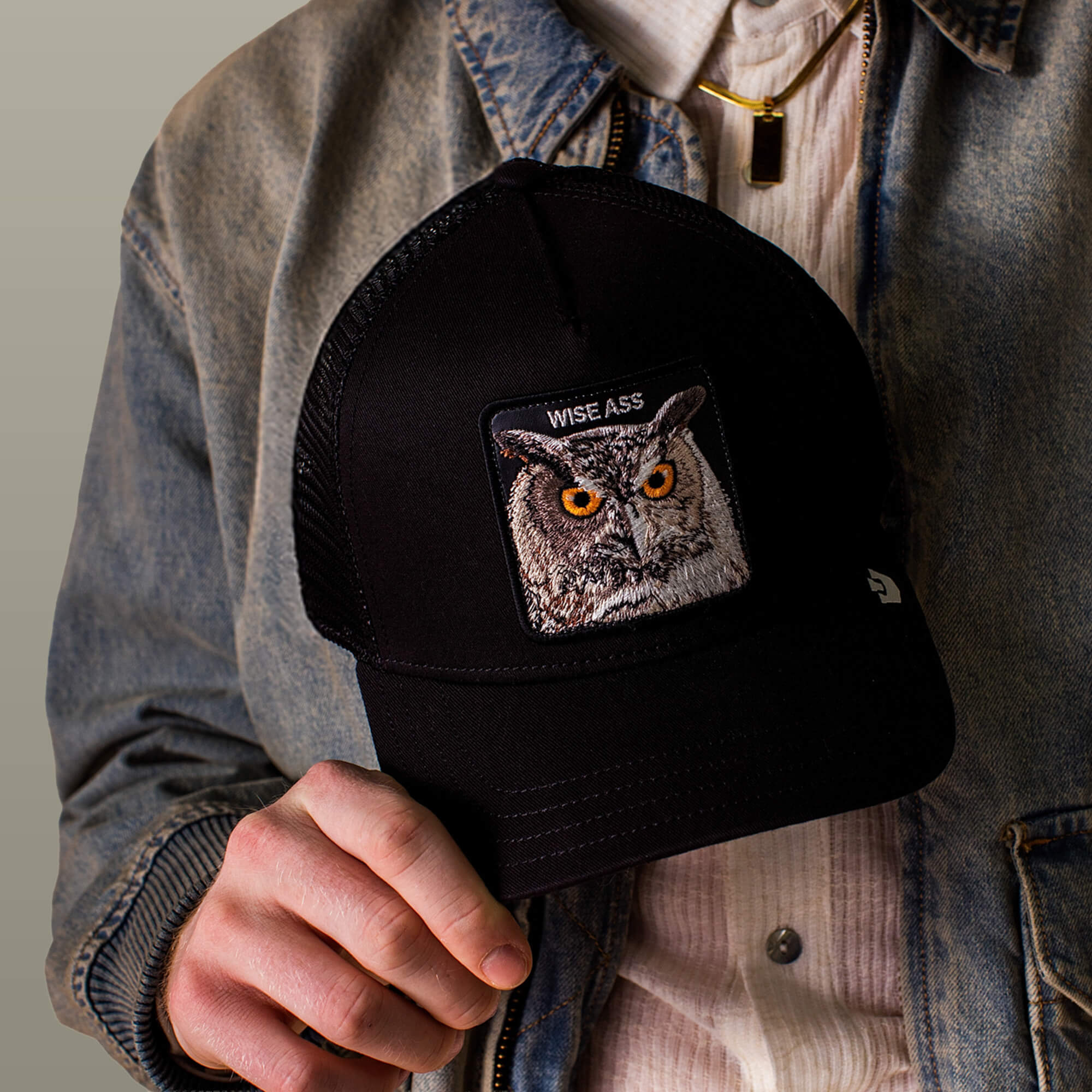 A person in a denim jacket holds the Wise Ass Trucker by Goorin Bros.—a black cap with an embroidered owl and "WISE ASS" text.