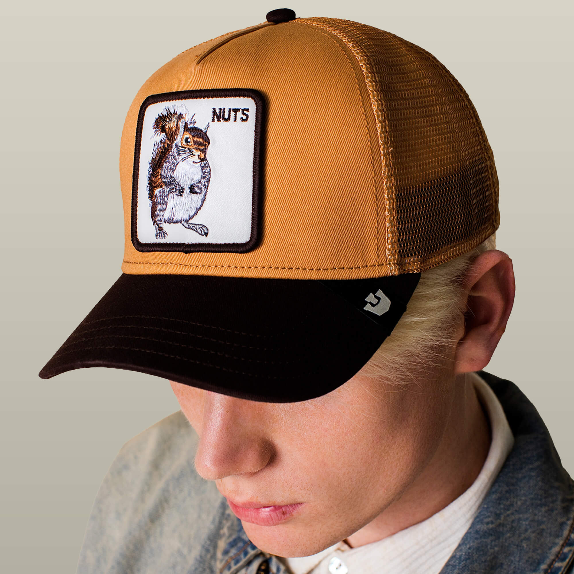 Person wearing the Goorin Bros. Nuts Trucker—a tan hat with a brown brim and a squirrel patch labeled "NUTS" on the front—paired with a light jacket against a neutral background.