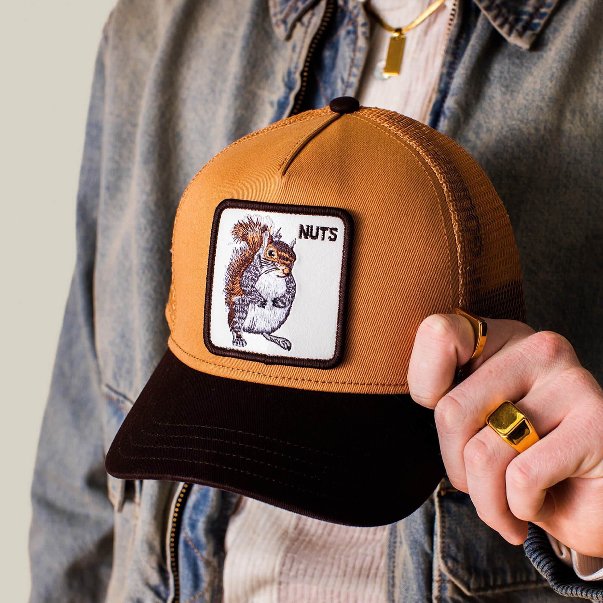 A person in a denim jacket holds the Goorin Bros. Nuts Trucker hat, which is tan with a brown brim and features a squirrel patch.