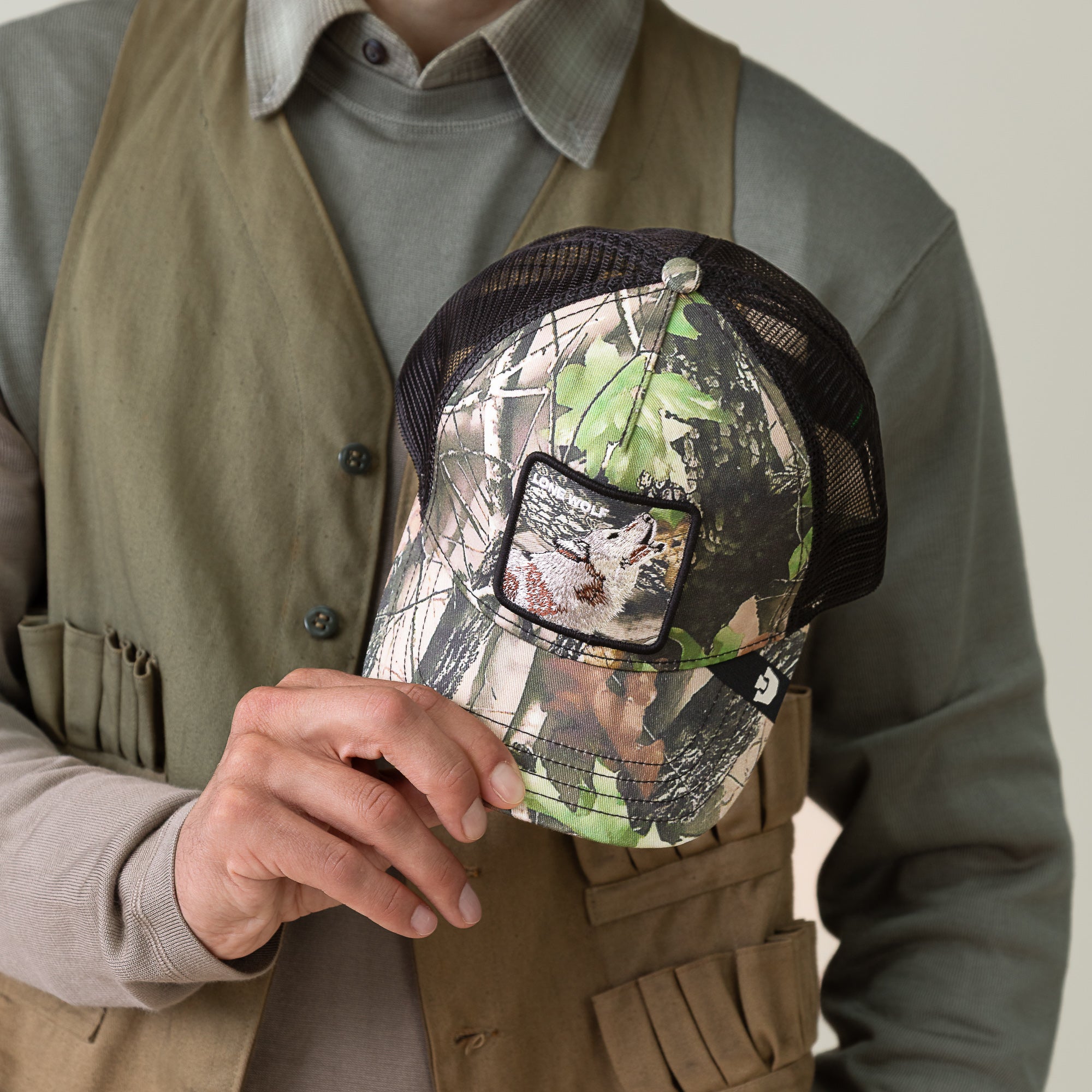 A person in a beige vest and gray shirt holds Goorin Bros.' Camouflage Lone Wolf cap, displaying its bold wolf patch during an off-grid soft launch.