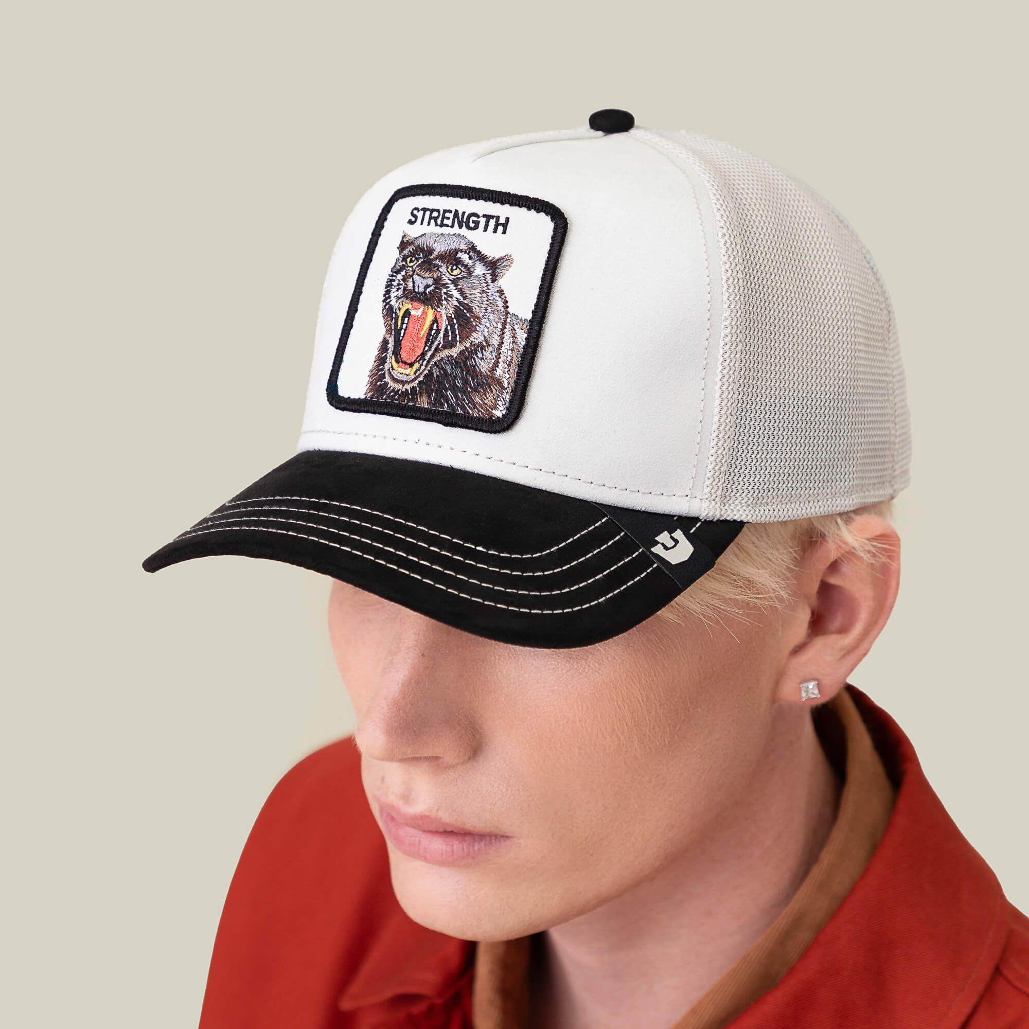A person wears the Goorin Bros. Suede Panther white crown baseball cap with a black brim, roaring panther patch, and STRENGTH label, paired with a red collared shirt and square stud earring against a plain background.