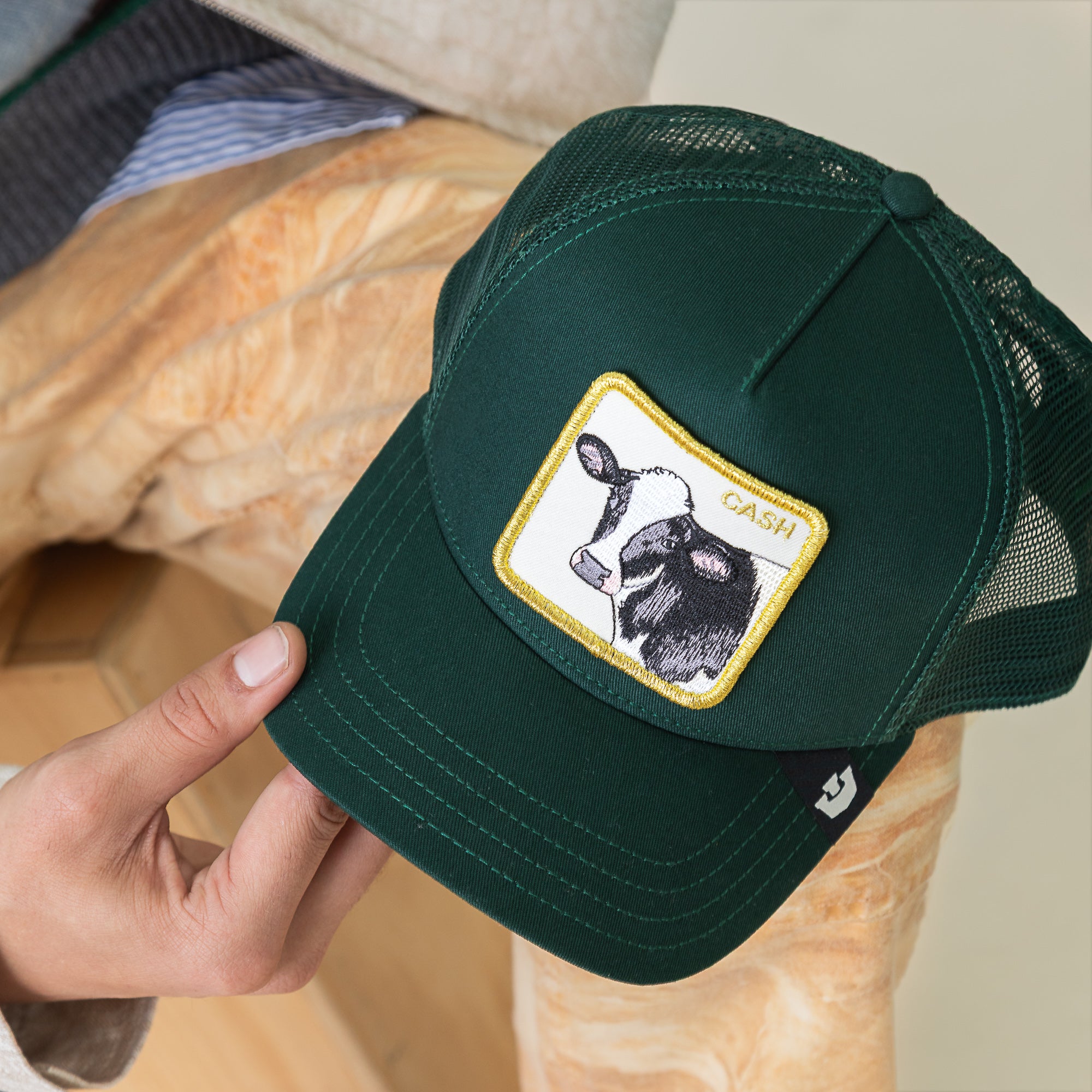A person holds the Goorin Bros. Cash Cow Trucker hat, a dark green cap featuring a bold bovine patch with "Cash Cow" on the front.